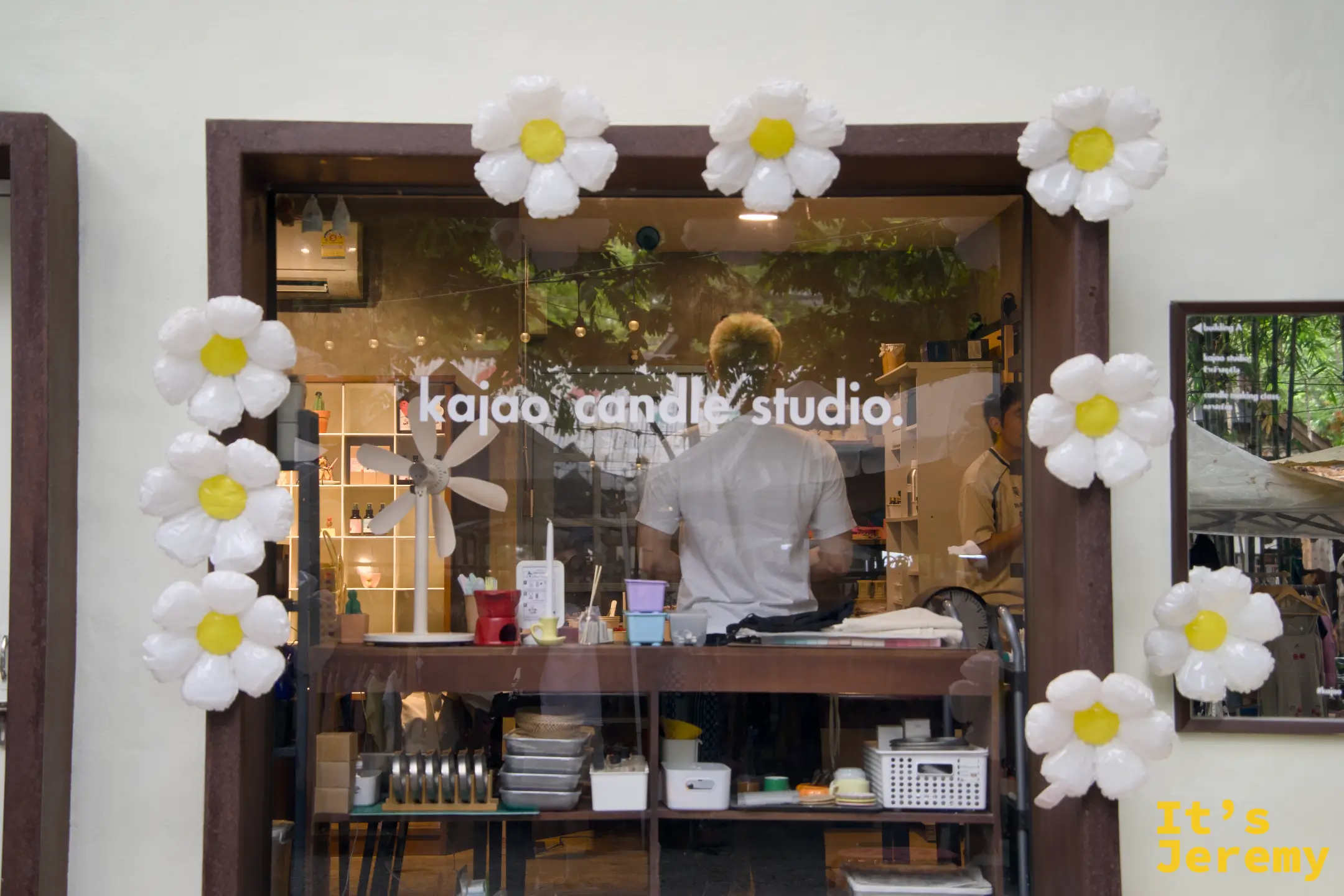 kajao candle studio 的工作室窗口，木窗框四周裝飾白色雛菊氣球，店員在裡面備料