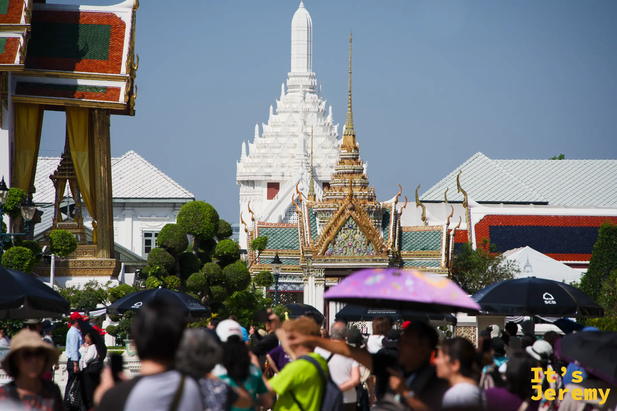 園區內擁擠的觀光人潮，遊客撐傘擋陽光，背景白色 Prasat 塔與金色屋頂