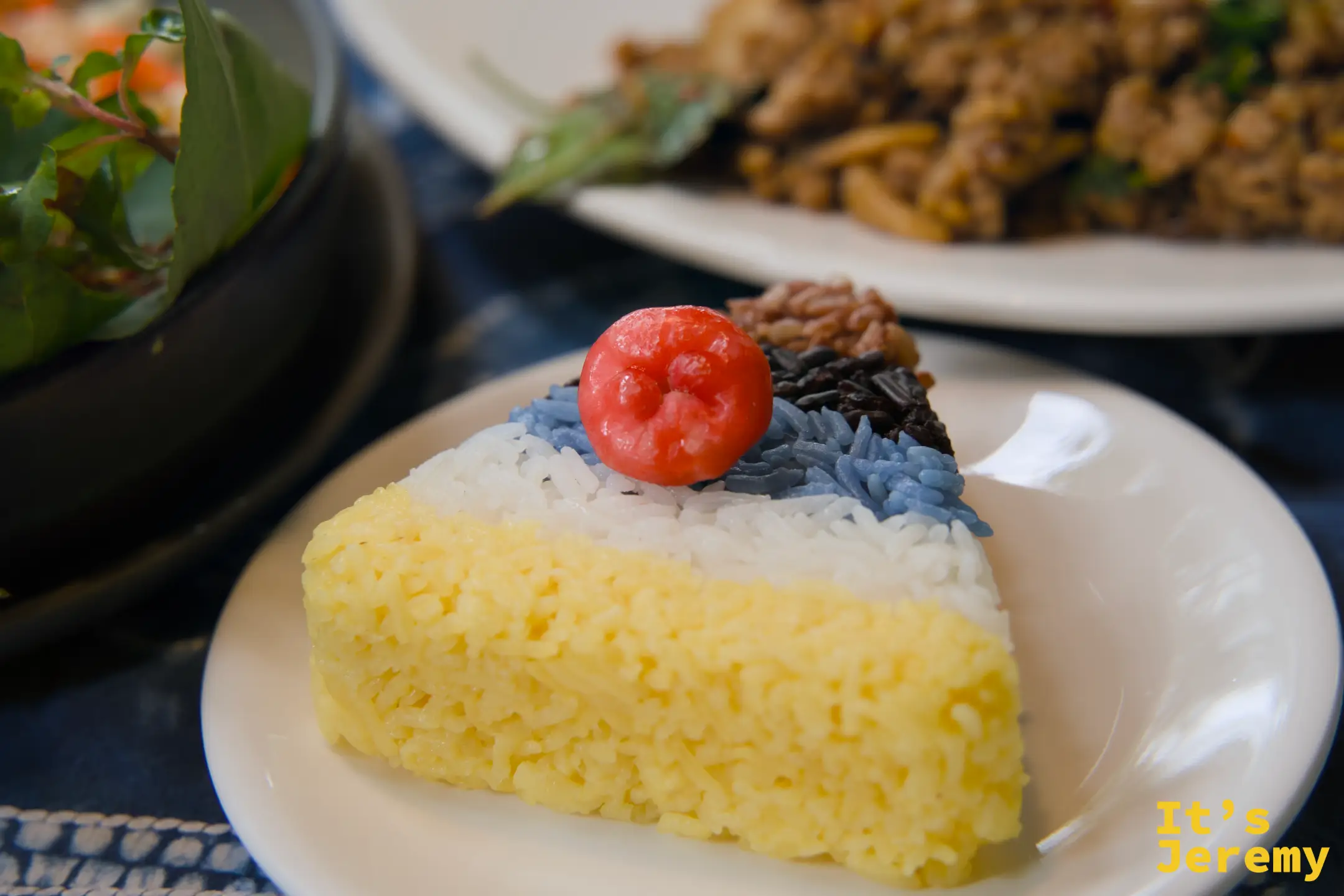 五色米飯特寫，三角形如蛋糕切片，底層薑黃色、中層白色、頂層藍紫黑三色，頂端放著一顆紅色小球