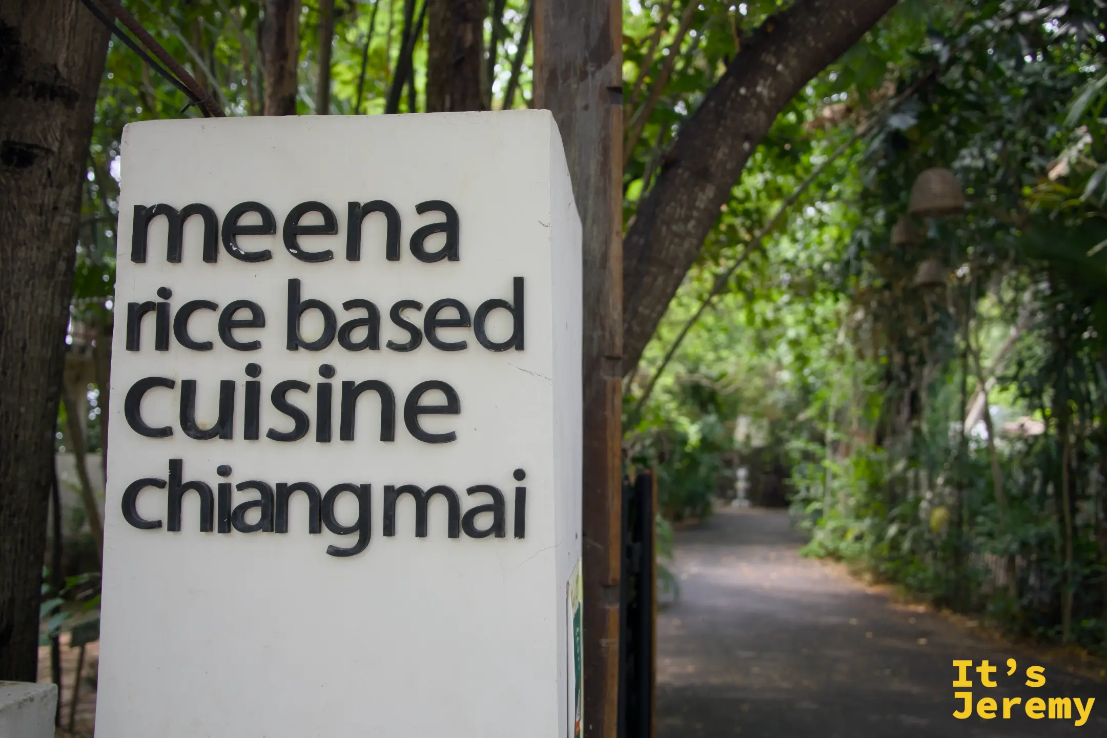 白色方柱招牌寫著 meena rice based cuisine chiangmai，後方是綠蔭步道與掛著銅鈴的樹