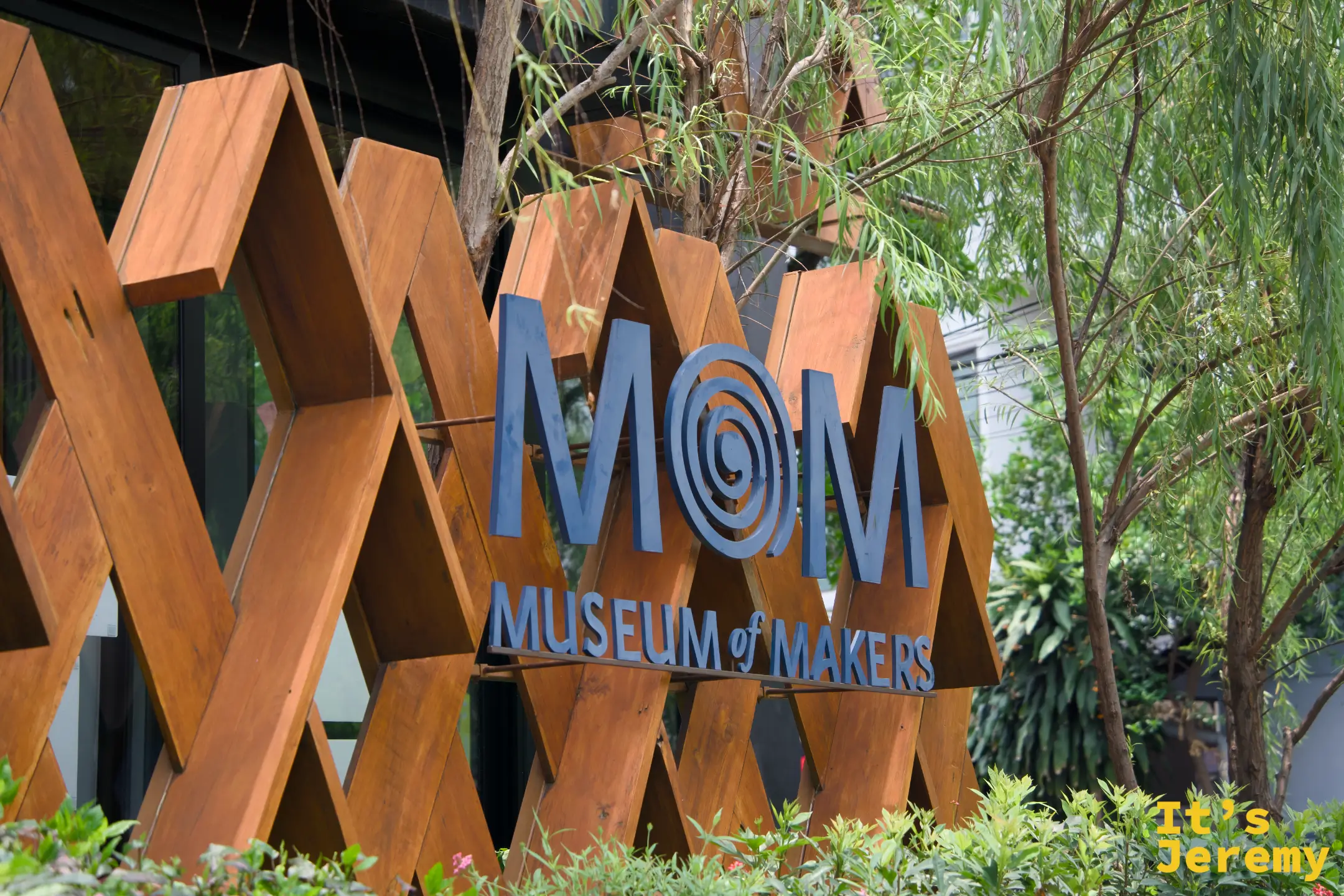 木格柵上的藍色 MOM（Museum of Makers）立體字標誌，周圍被樹葉環繞