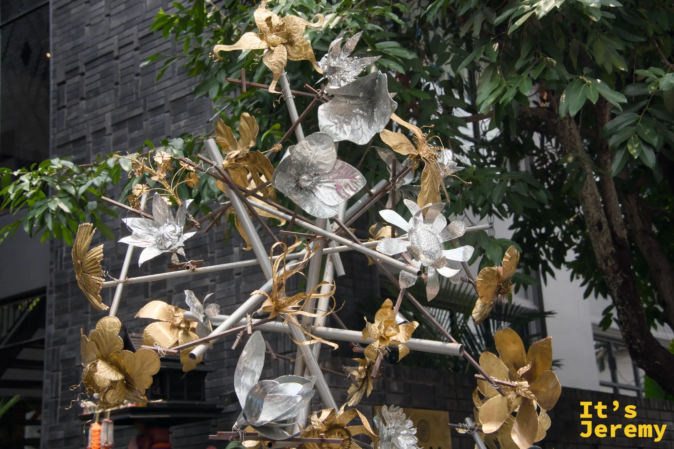 銀色與金色金屬花朵組成的雕塑，立在灰磚建築前的綠樹之間