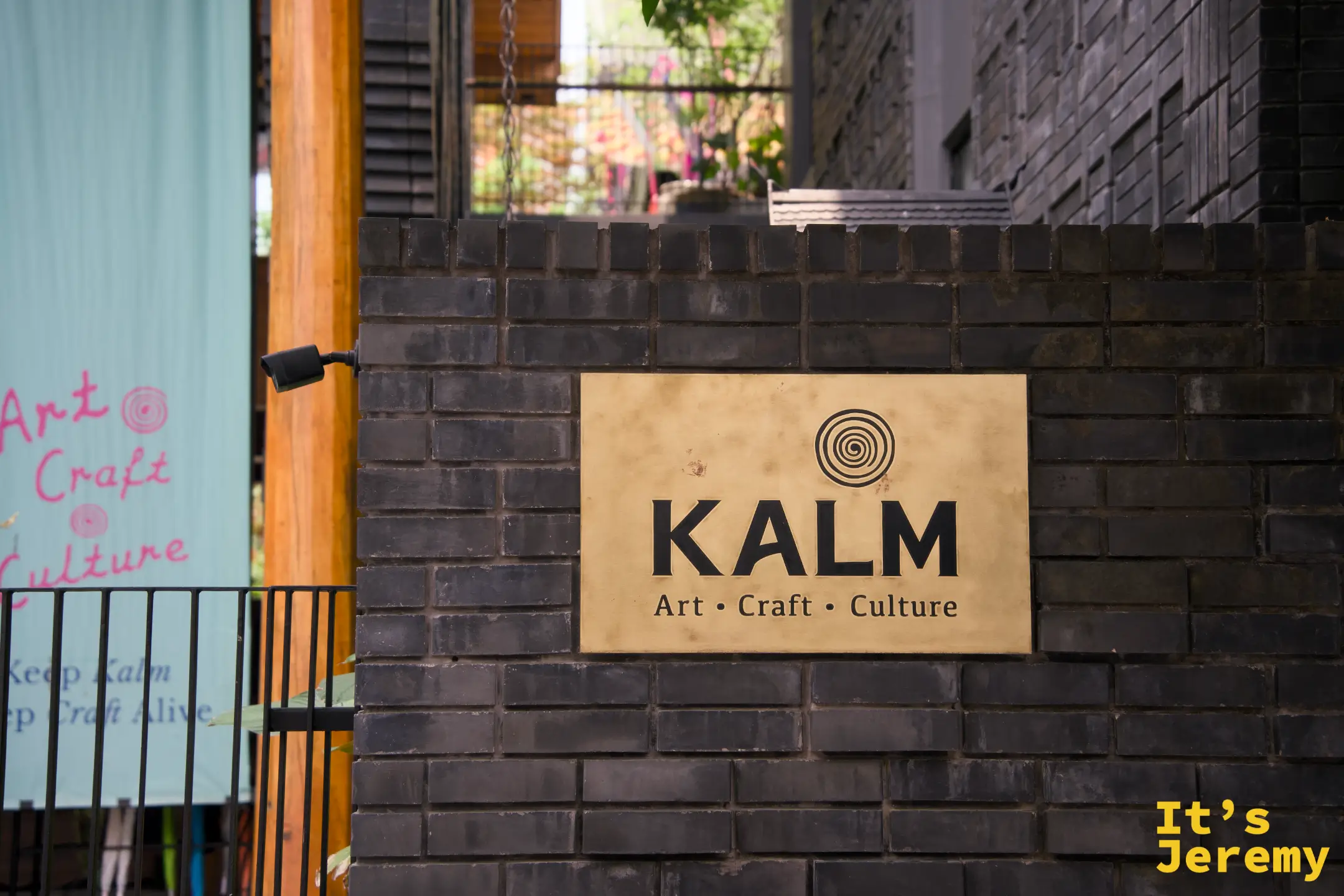 灰磚牆上的金色 KALM 銅牌，寫著 Art・Craft・Culture，左後方是湖水綠的「Keep Kalm, Keep Craft Alive」布旗