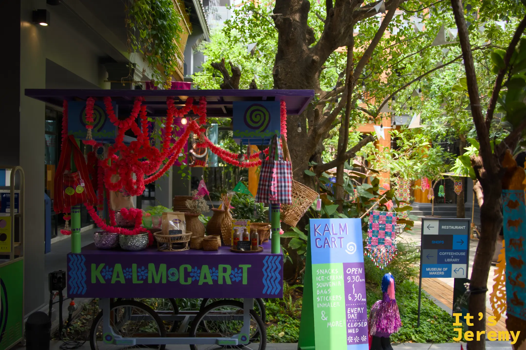 紫色的 KALM CART 小攤車，掛滿橘色花環與藤編籃，停在中庭樹下
