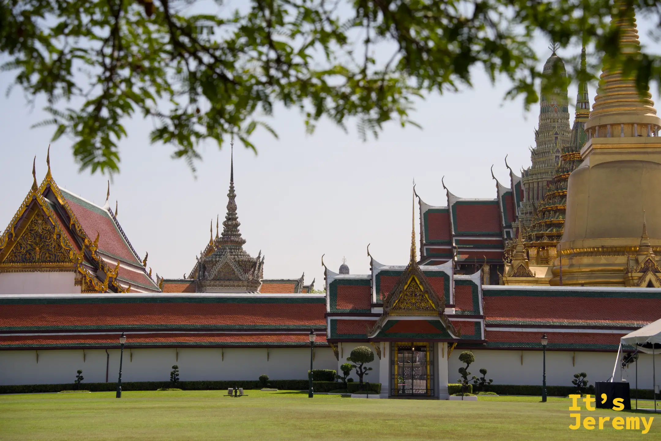 大皇宮外牆全景，樹蔭前景下的金色 Chedi 與紅綠瓦屋頂