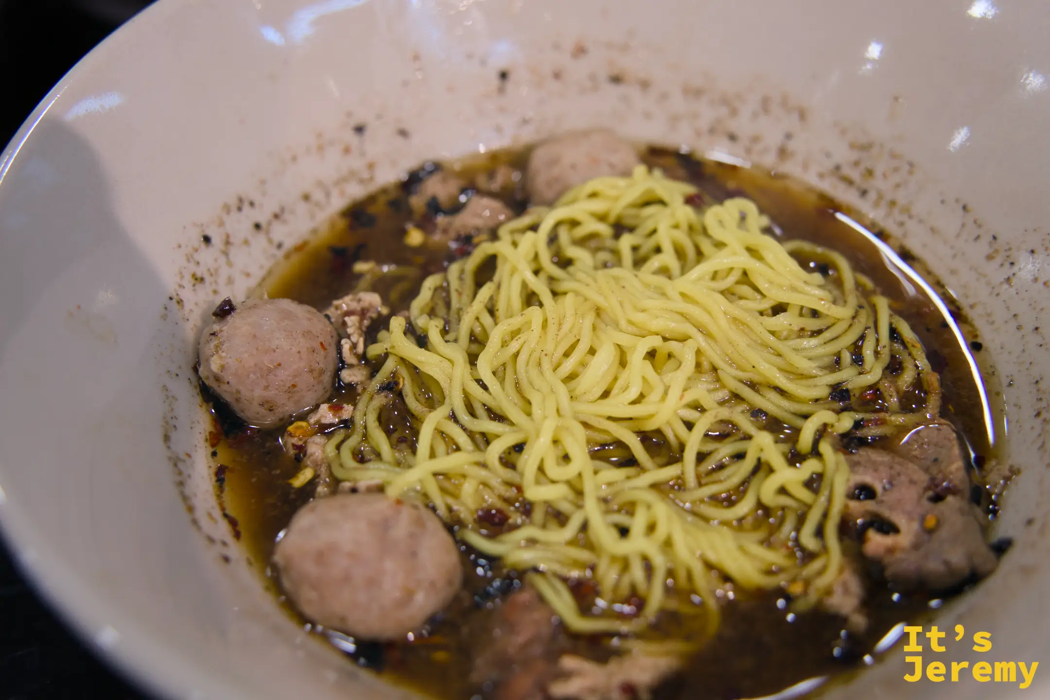 白碗裡的牛肉船麵，黃色雞蛋麵浸在深色辣湯中，湯裡有多顆牛肉丸與肉末，碗緣沾著辣椒碎
