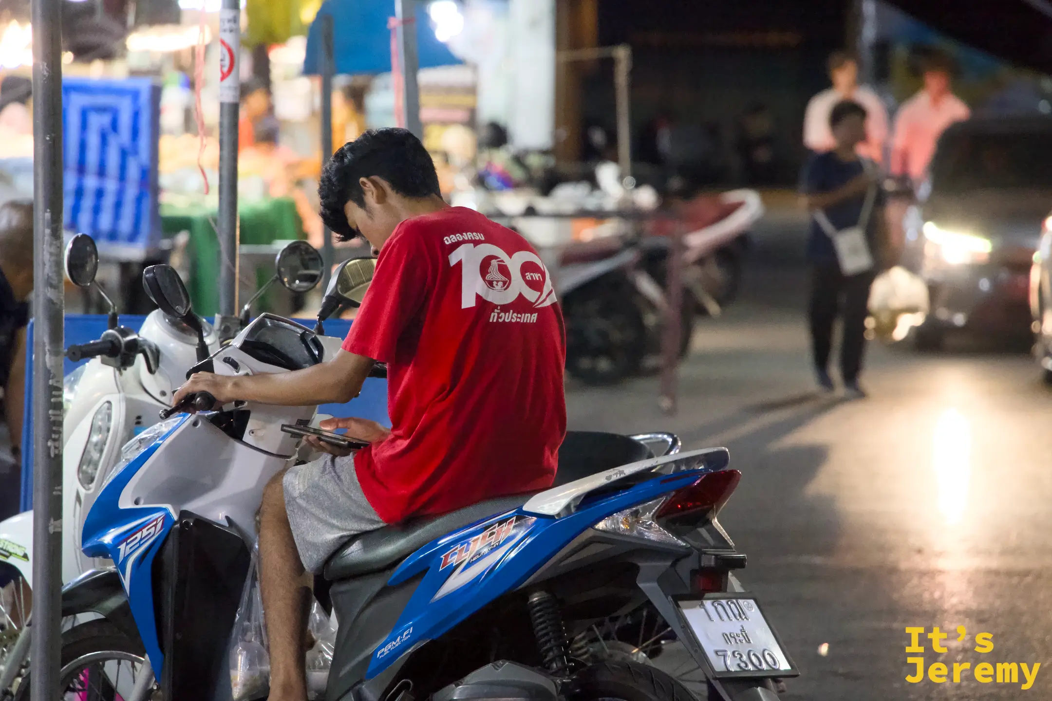 穿紅色上衣的少年跨坐在藍白色機車上低頭滑手機，背景是燈火通明的夜市攤位與來往人群