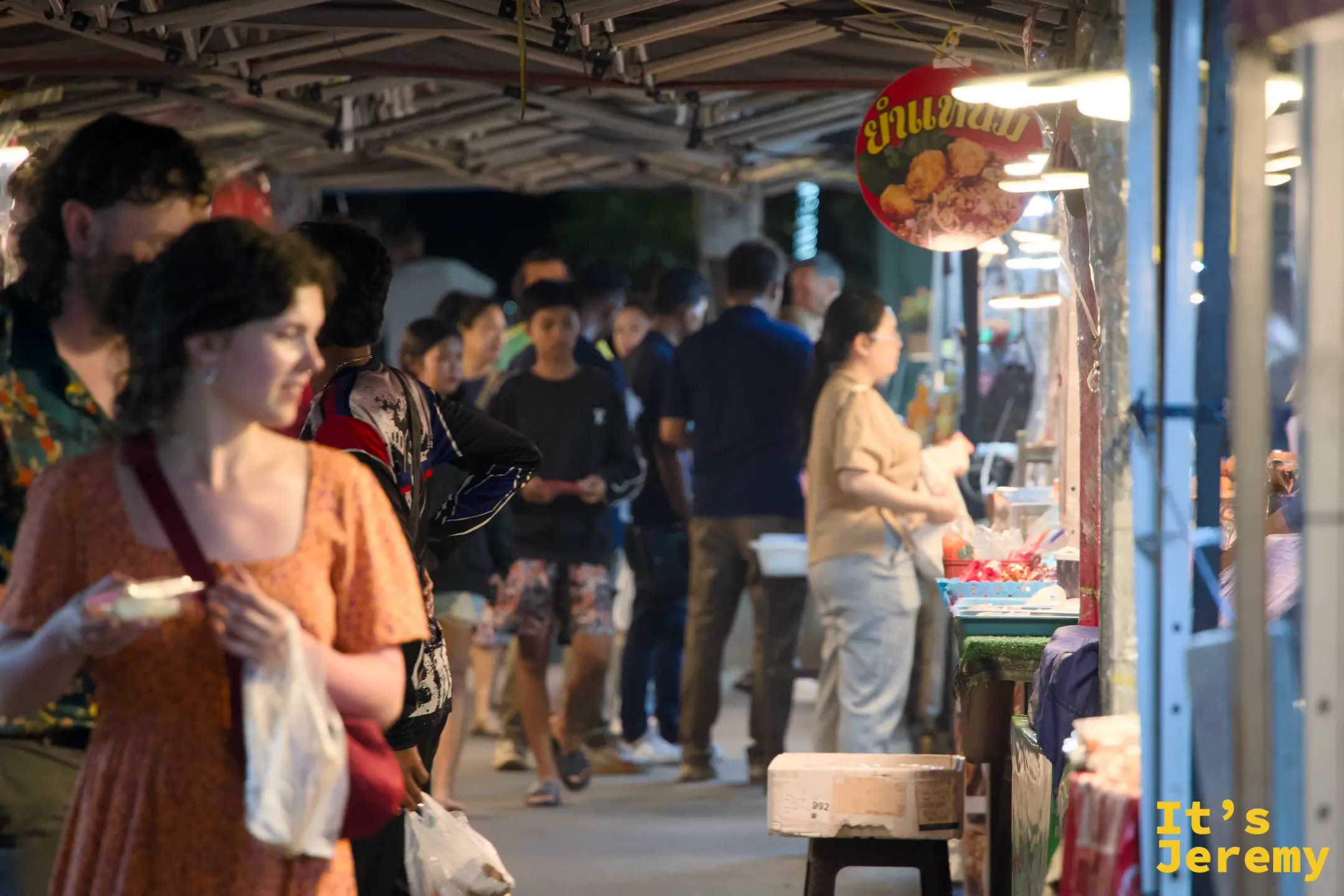 夜市帳棚下的走道擠滿人潮，前景一對外國旅人正端著小吃邊走邊吃，右側攤位掛著泰文圓形招牌
