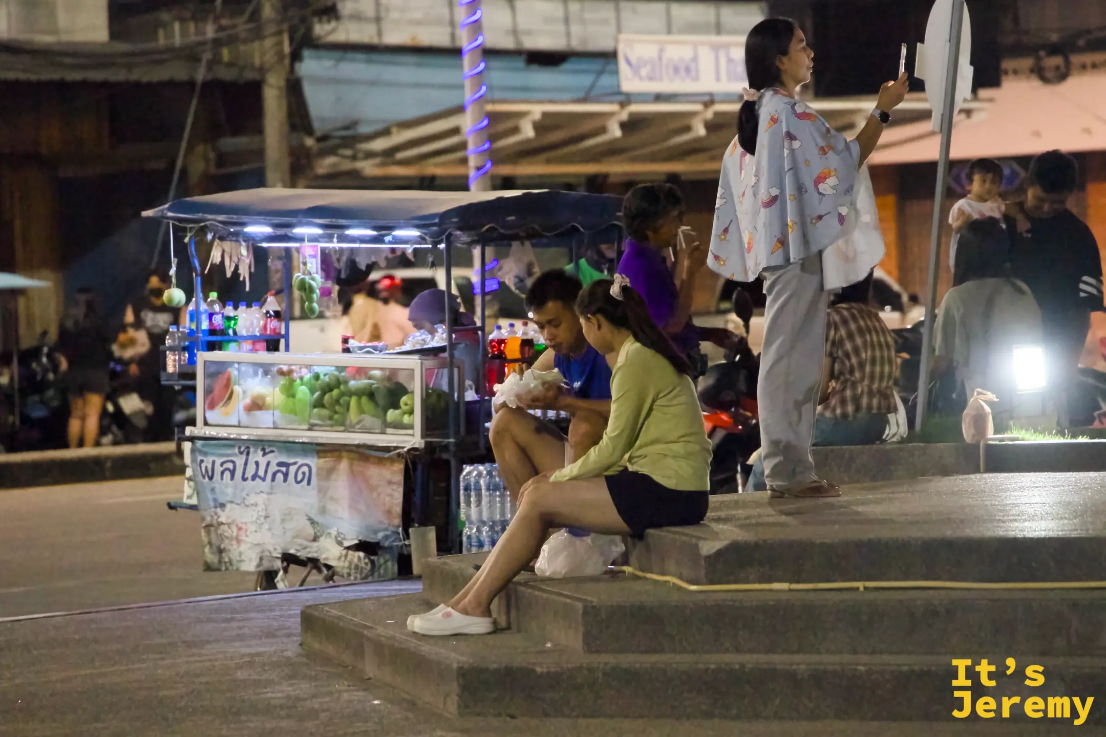 寫著泰文「新鮮水果」布條的水果攤前，一對年輕男女坐在階梯上分食袋裝水果