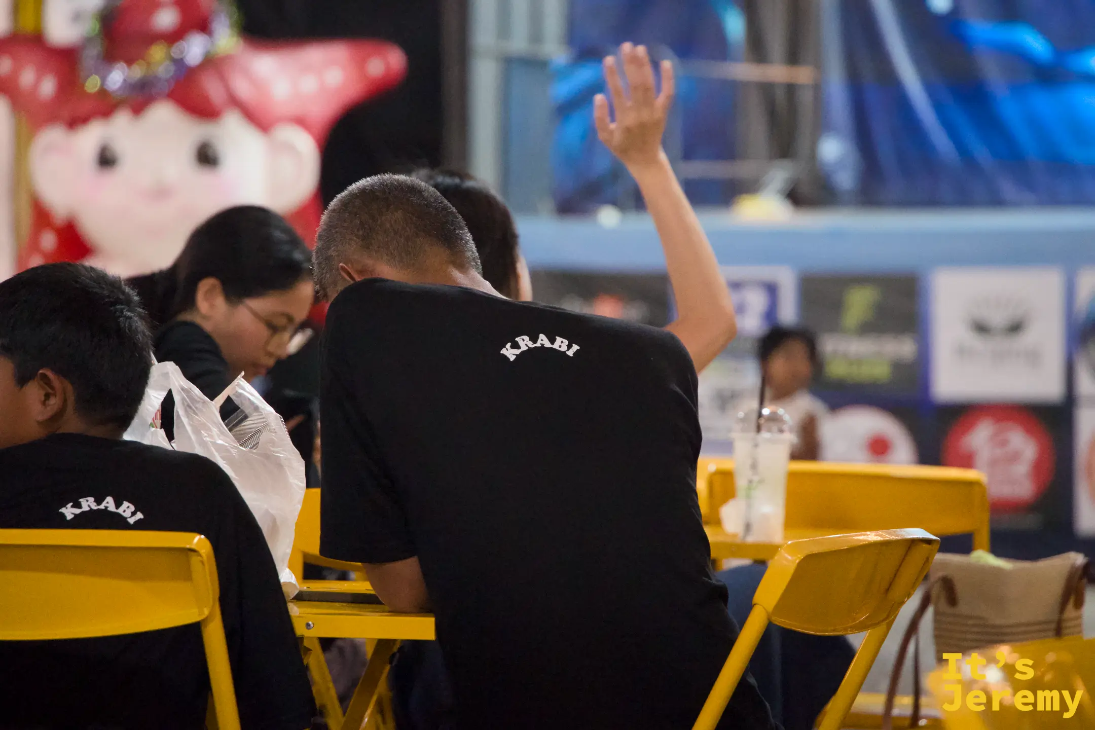 用餐區黃色桌椅旁坐著數名穿黑色 KRABI 字樣 T 恤的遊客，背景有一隻紅色海星造型的人偶裝飾