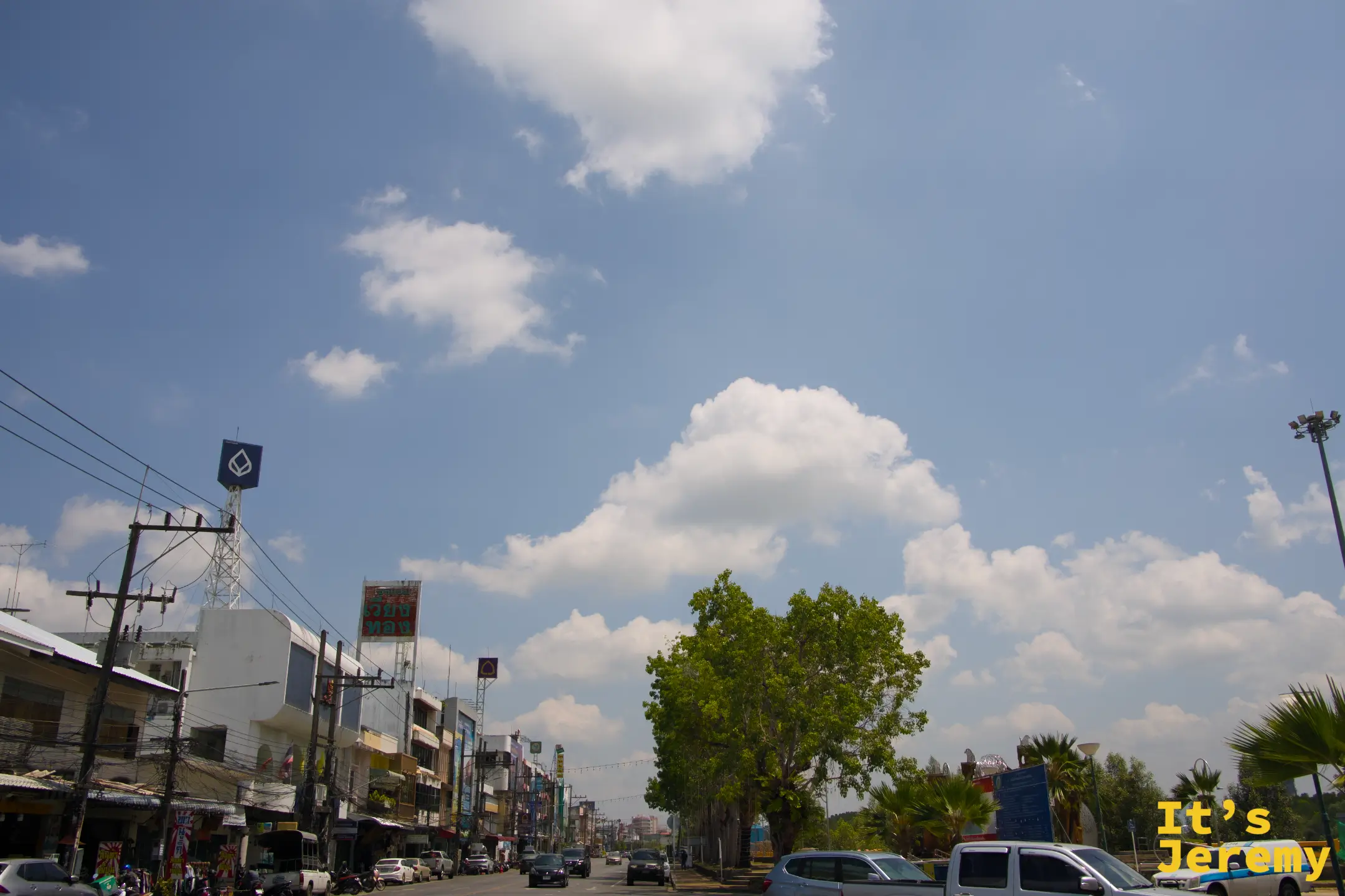 喀比鎮市區主幹道，藍天白雲、兩側商店招牌排開，一棵大樹矗立路中央
