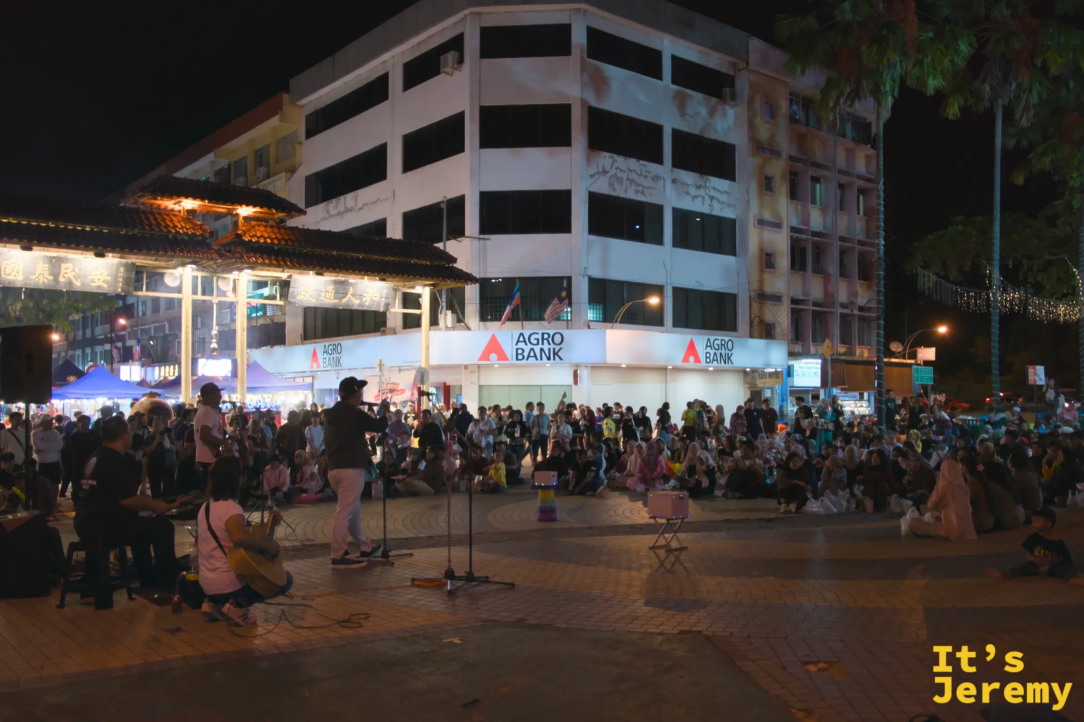 一支樂隊在加雅街廣場夜晚表演，數百名觀眾圍坐在前方，背景是 Agro Bank 建築