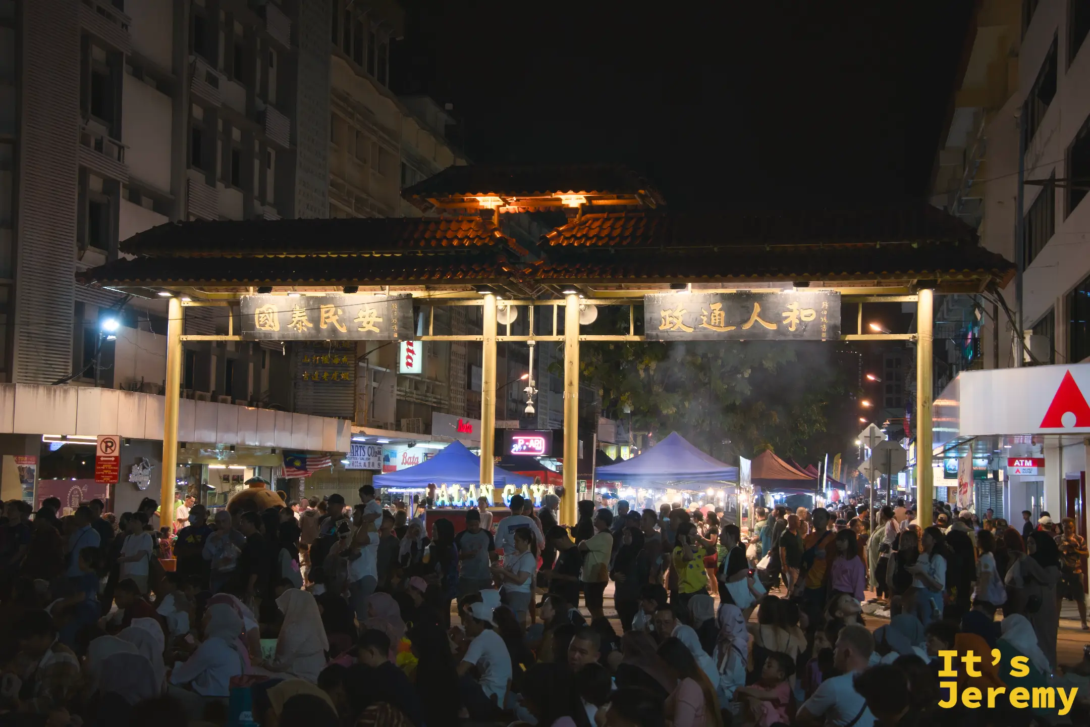 加雅街入口的中式牌坊在夜晚燈光照耀下，牌坊下方擠滿了人群，遠處可見藍色帳篷攤位