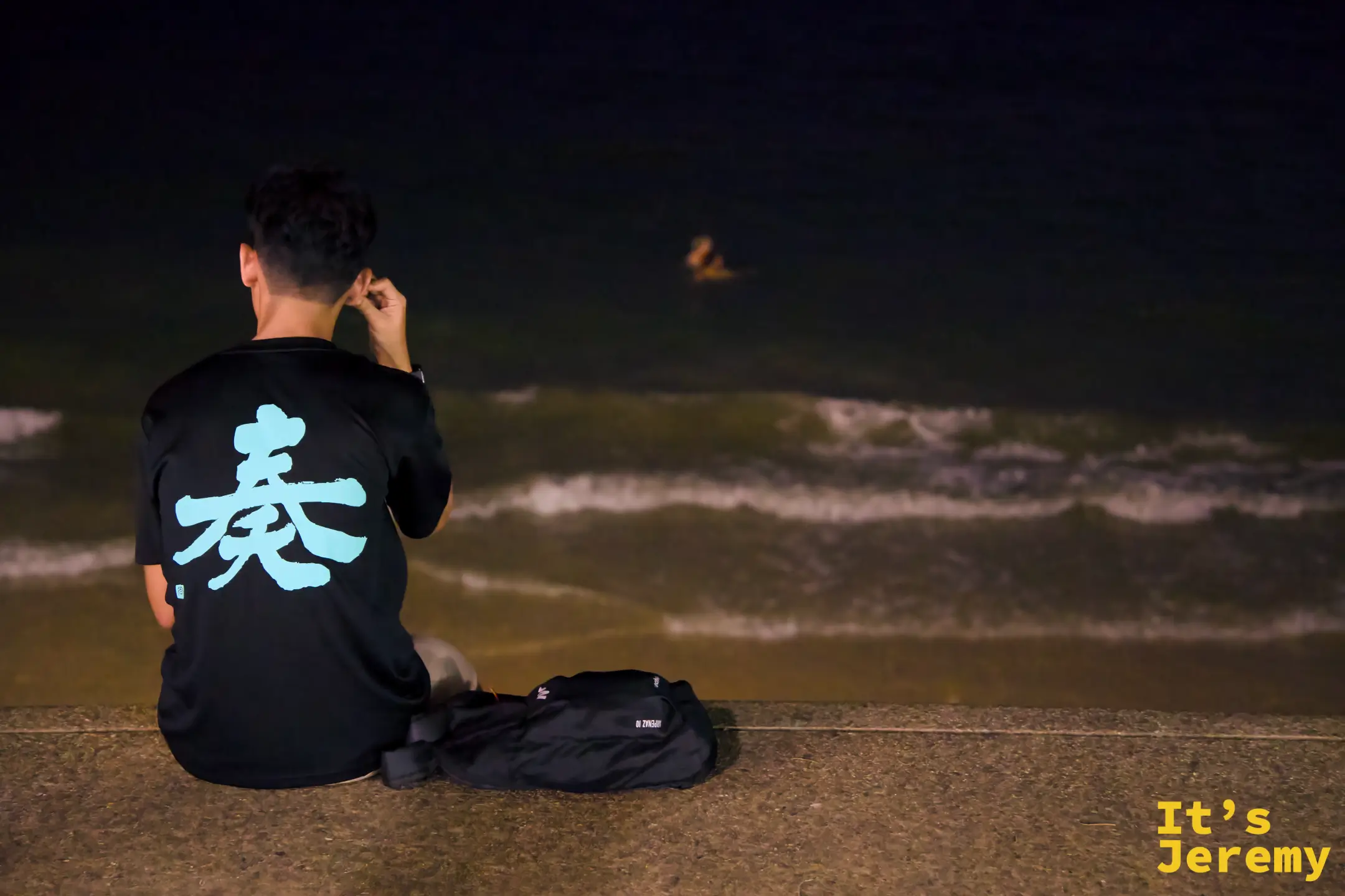 入夜後一名旅人背對鏡頭坐在低矮圍牆邊，望向深黑色海面，遠處有人在夜間游泳