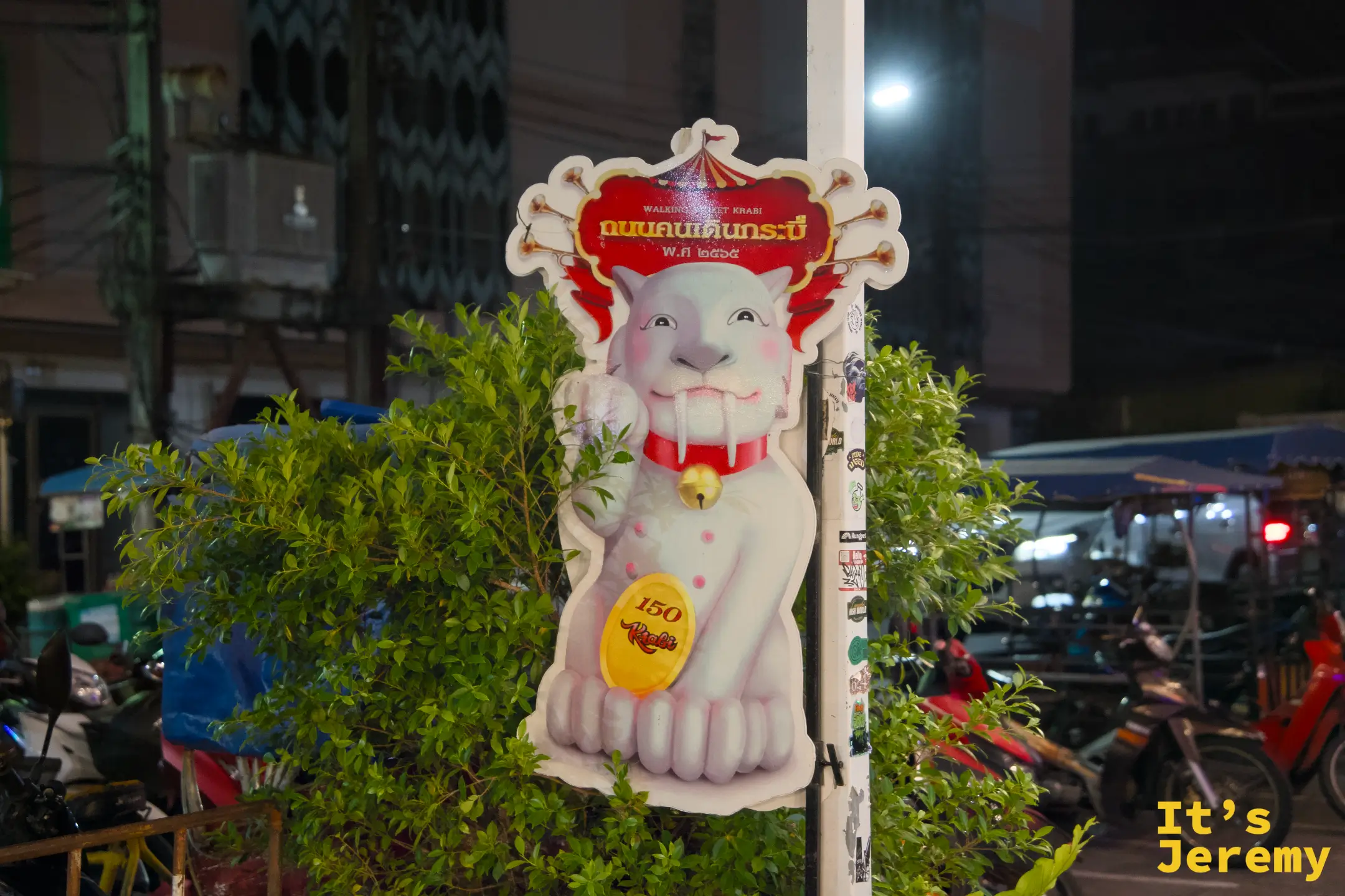 喀比鎮夜晚路口的 Walking Street Krabi 指示立牌，白色惡魔貓咪圖案立在植物叢前，背景是摩托車與夜市燈光