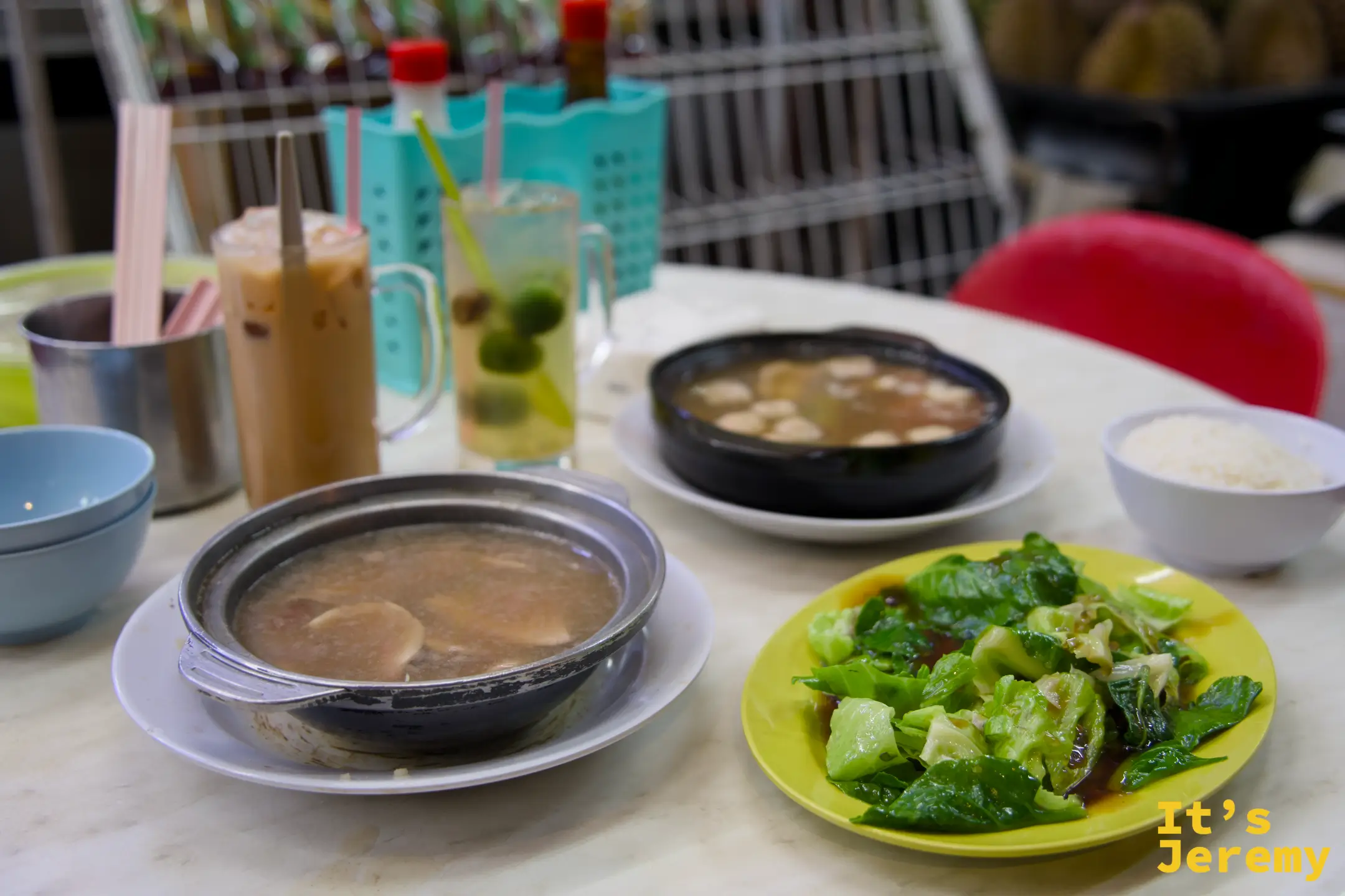 全桌擺設：銀色黃酒肝湯、黑砂鍋肉骨茶、黃盤蔬菜、奶茶和柑橘飲料，右後方可見等著被切的榴槤