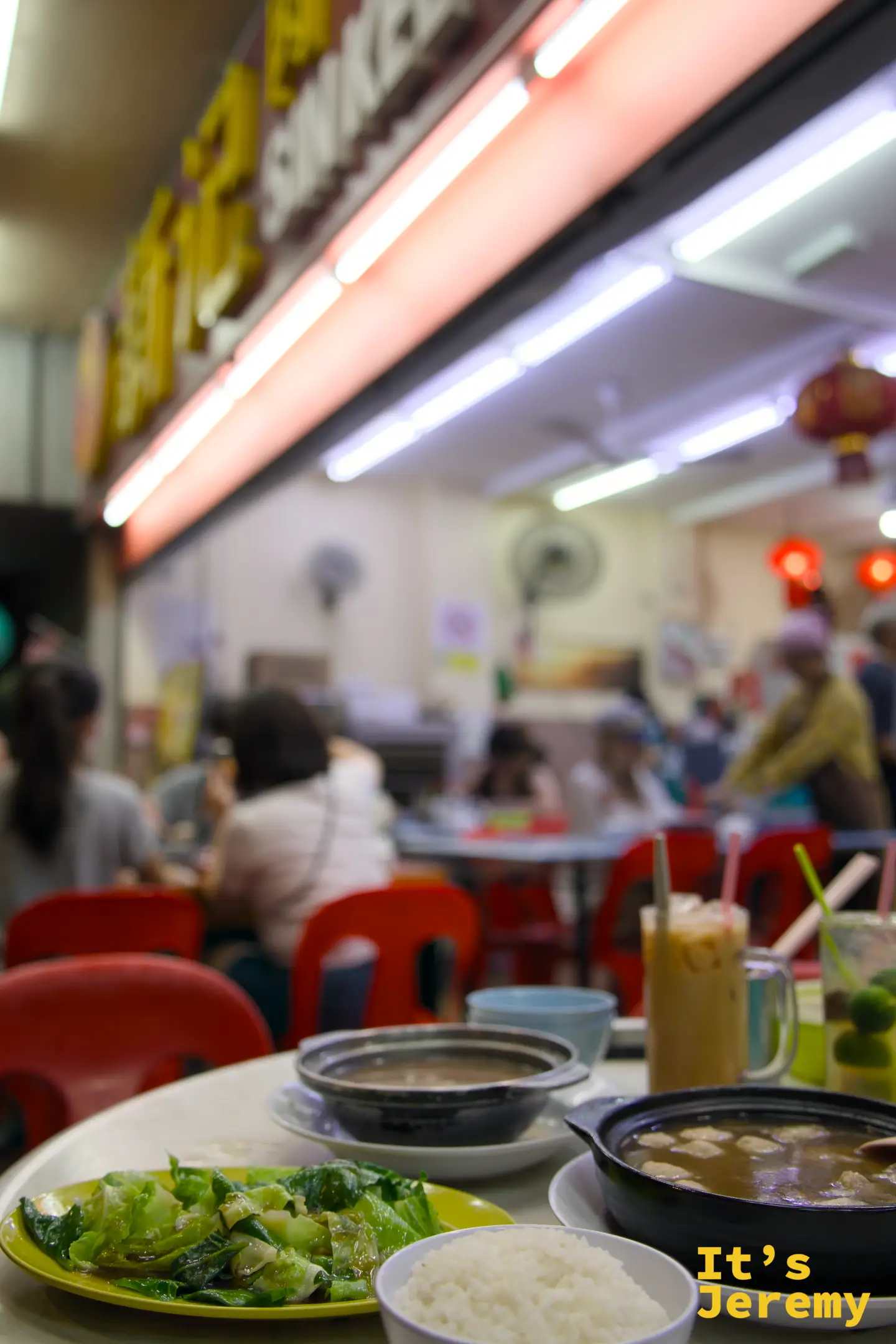 新記肉骨茶桌面近景：黑色砂鍋、炒青菜、白米飯、冰奶茶與青檸飲料排開，後方另一砂鍋模糊