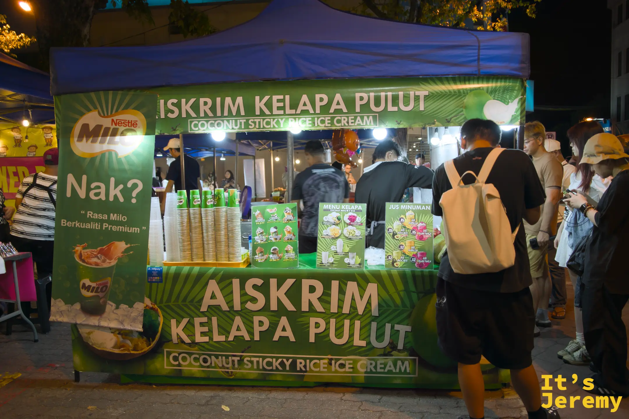 Iskrim Kelapa Pulut 椰子糯米冰淇淋攤位外觀，藍色帳篷搭配 Milo 品牌綠色招牌，多名顧客在旁研究菜單