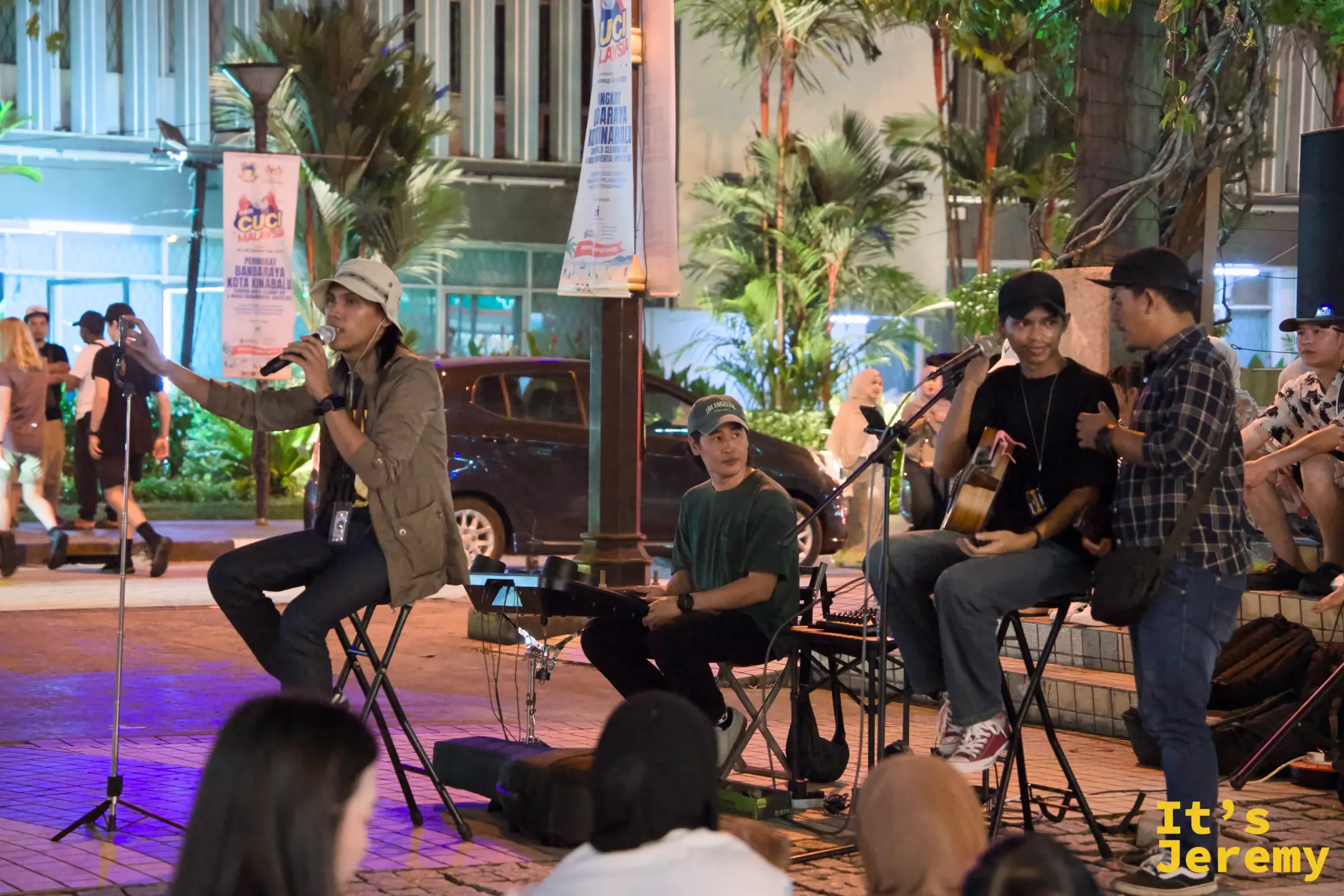 一支小型樂隊在廣場前的夜晚演出，歌手坐著演唱，旁邊有吉他手與鍵盤手