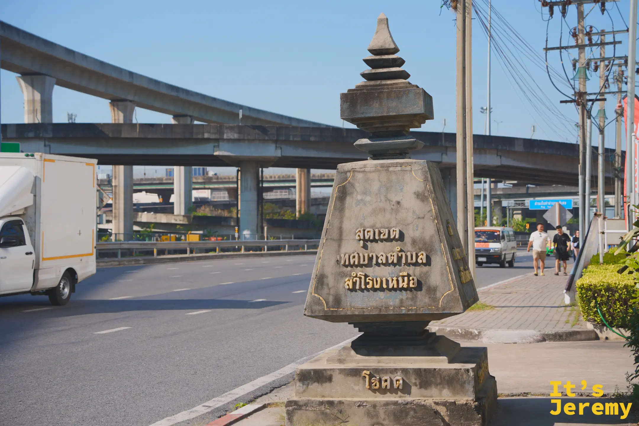 馬路邊的「Samrong Nuea 市鎮邊界」傳統石碑，背景是 BTS 高架軌道