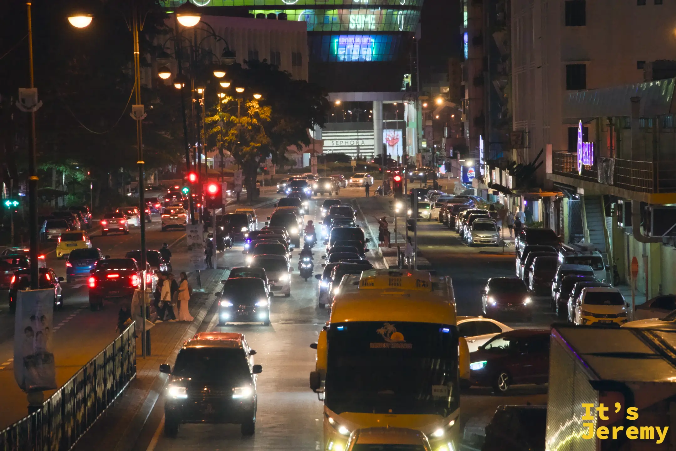 亞庇市區夜晚主幹道，車流繁忙，街底可見 Sephora 購物商場大型廣告燈，路燈連成一線