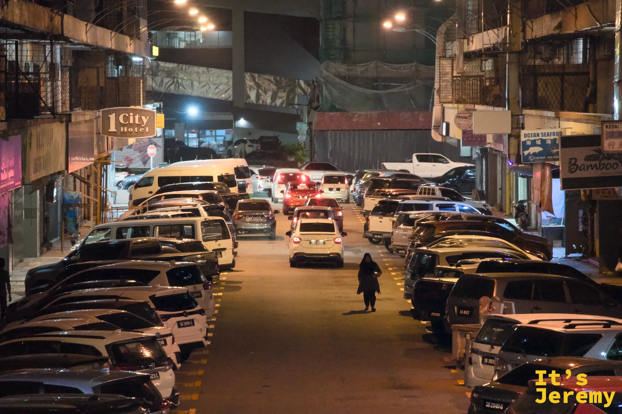 亞庇停車場巷弄夜景，1City Hotel 招牌與 Ocean Seafood Bamboo 餐廳招牌分列兩側，停滿車輛