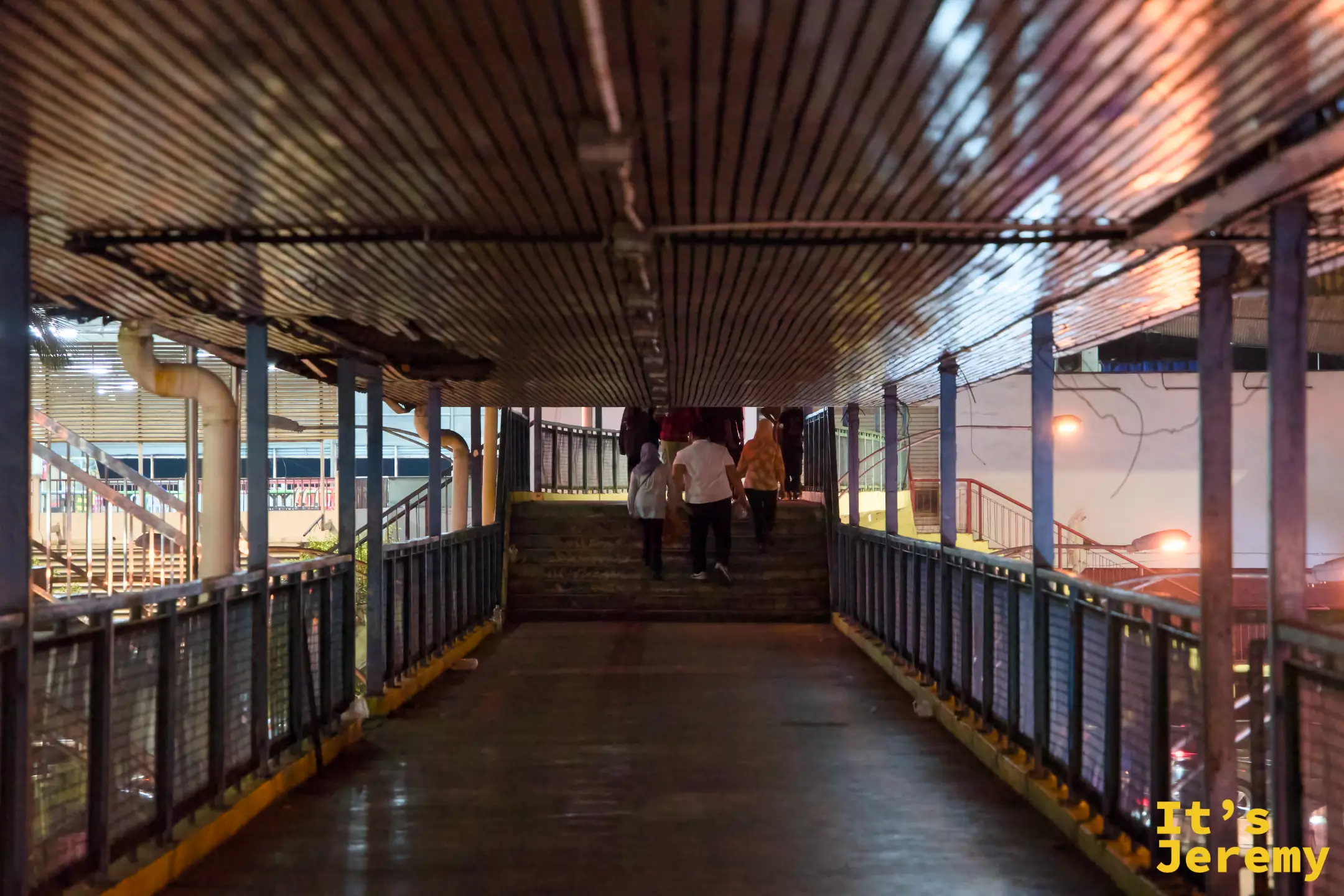 夜間行人天橋走道，幾人走向遠處樓梯，天橋頂為波浪形金屬板，燈光昏黃