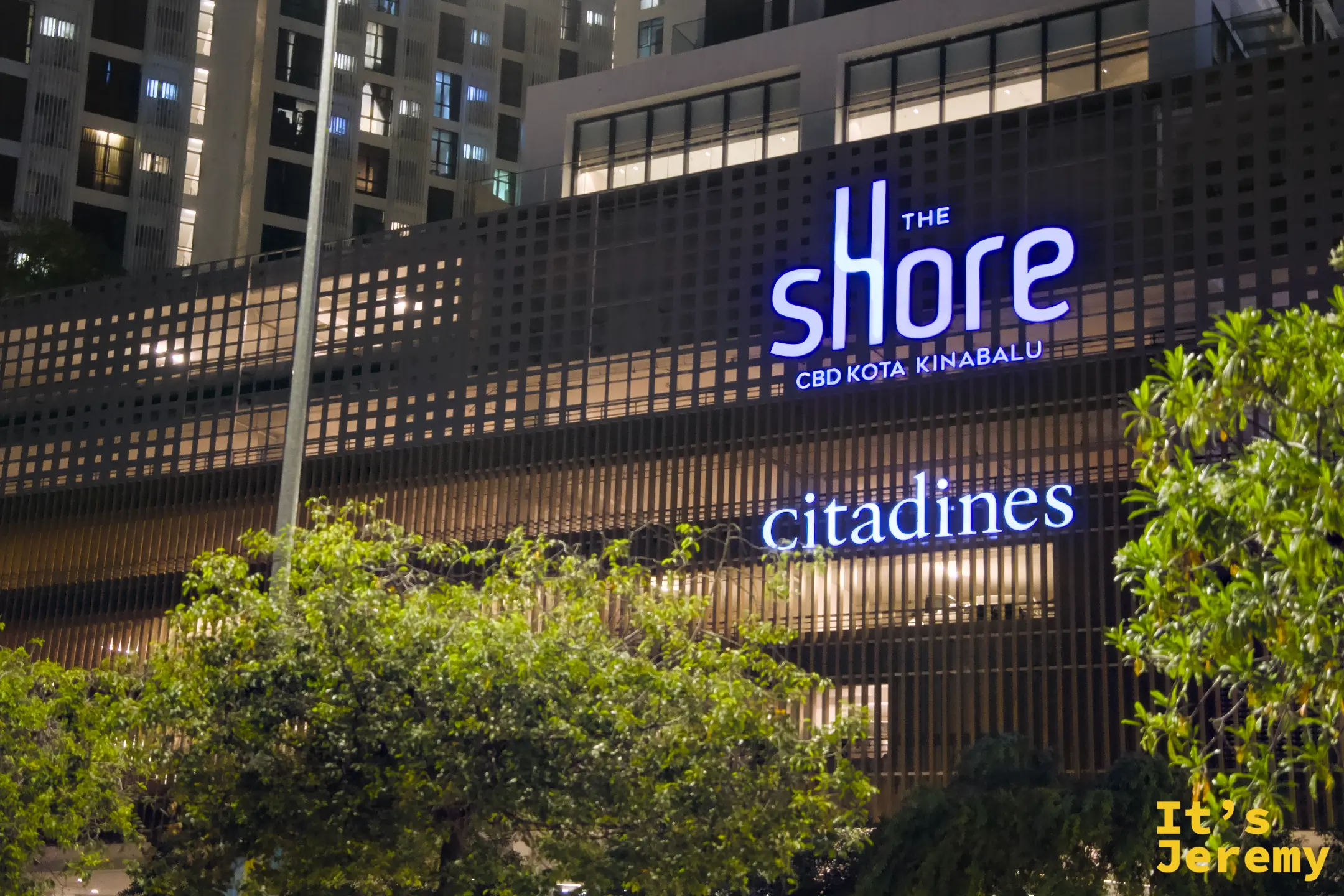 The Shore CBD Kota Kinabalu 與 Citadines 飯店夜間招牌，藍色霓虹字體亮起，前景為綠色樹叢