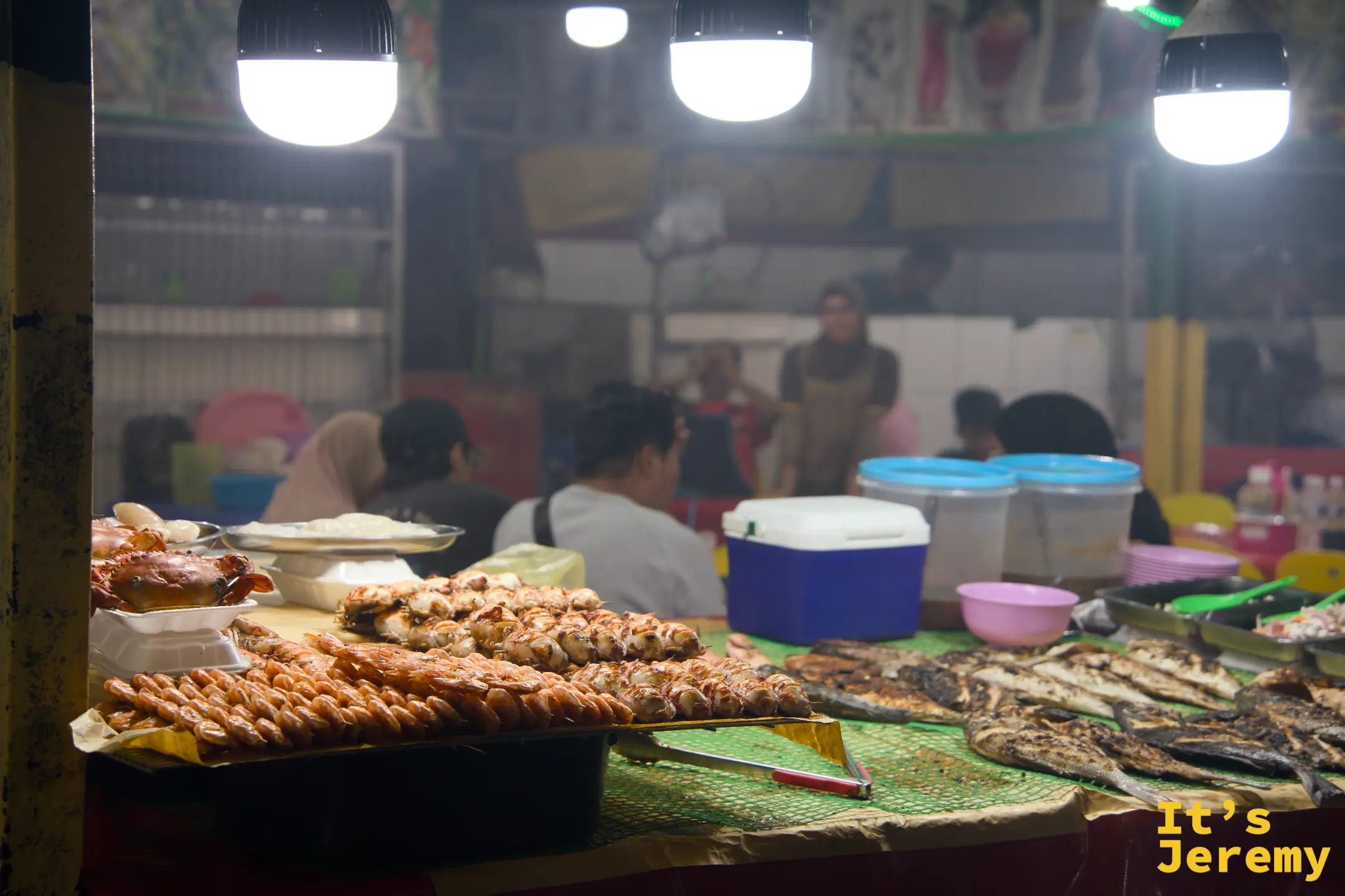 夜市海鮮攤前的陳列：螃蟹、蝦子竹串與整尾烤魚並排展示，燈光打亮了每一道蝦殼