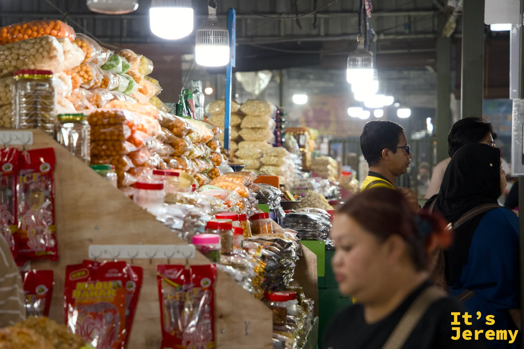 乾貨走道：顧客沿著掛滿乾貨和零食的走廊慢慢選購，前方有一名戴頭巾的女顧客正在看貨品