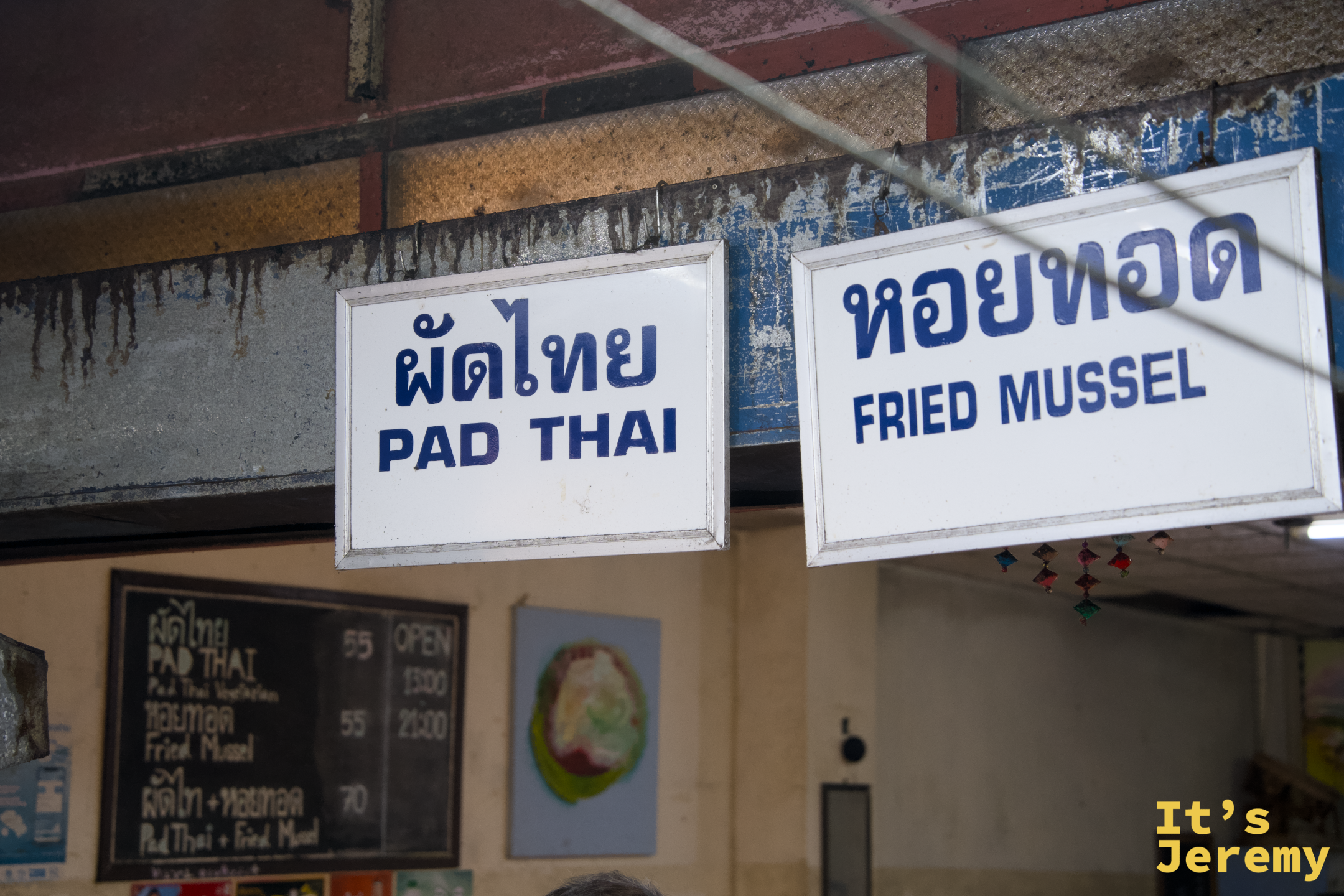 掛牌招牌上寫著 PAD THAI 與 FRIED MUSSEL，藍底白字，掛在老攤棚架上