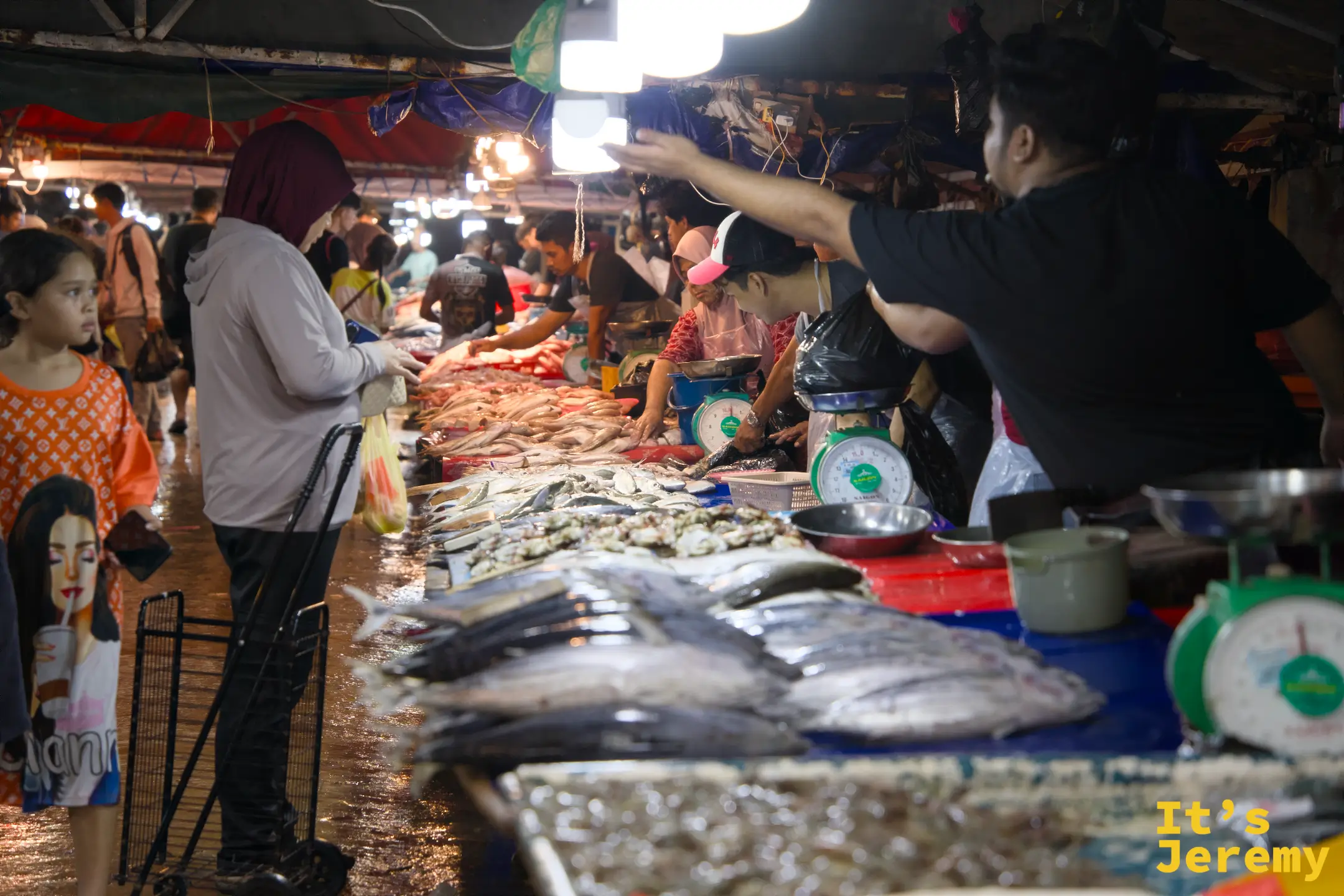 生鮮魚市走廊：左邊是帶著小孩的女顧客正在挑魚，右邊多名攤主在攤位後忙著秤重切魚，兩台磅秤同時運作
