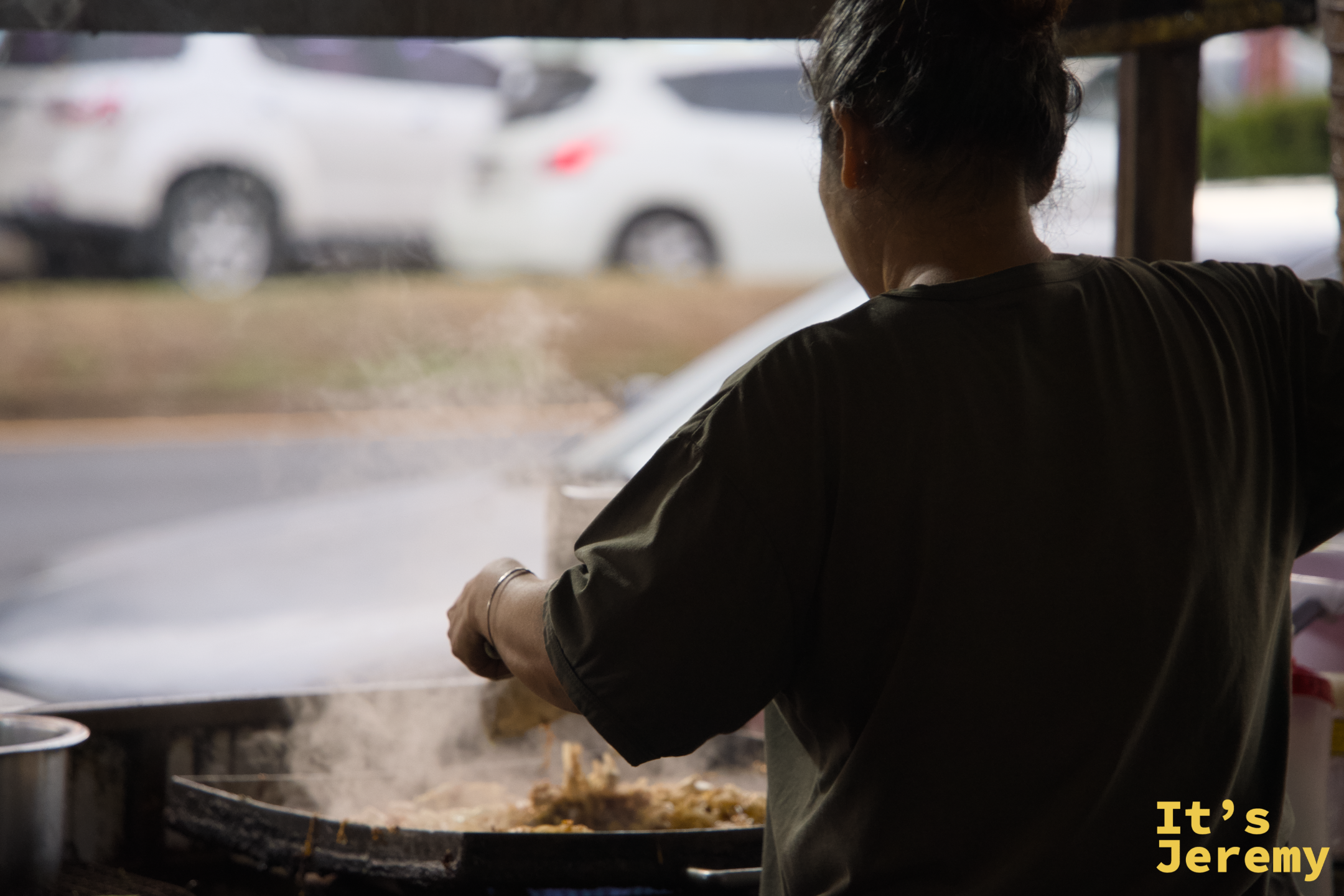 師傅背對鏡頭，全神貫注地在大圓鍋裡翻炒，白色油煙往棚外飄散