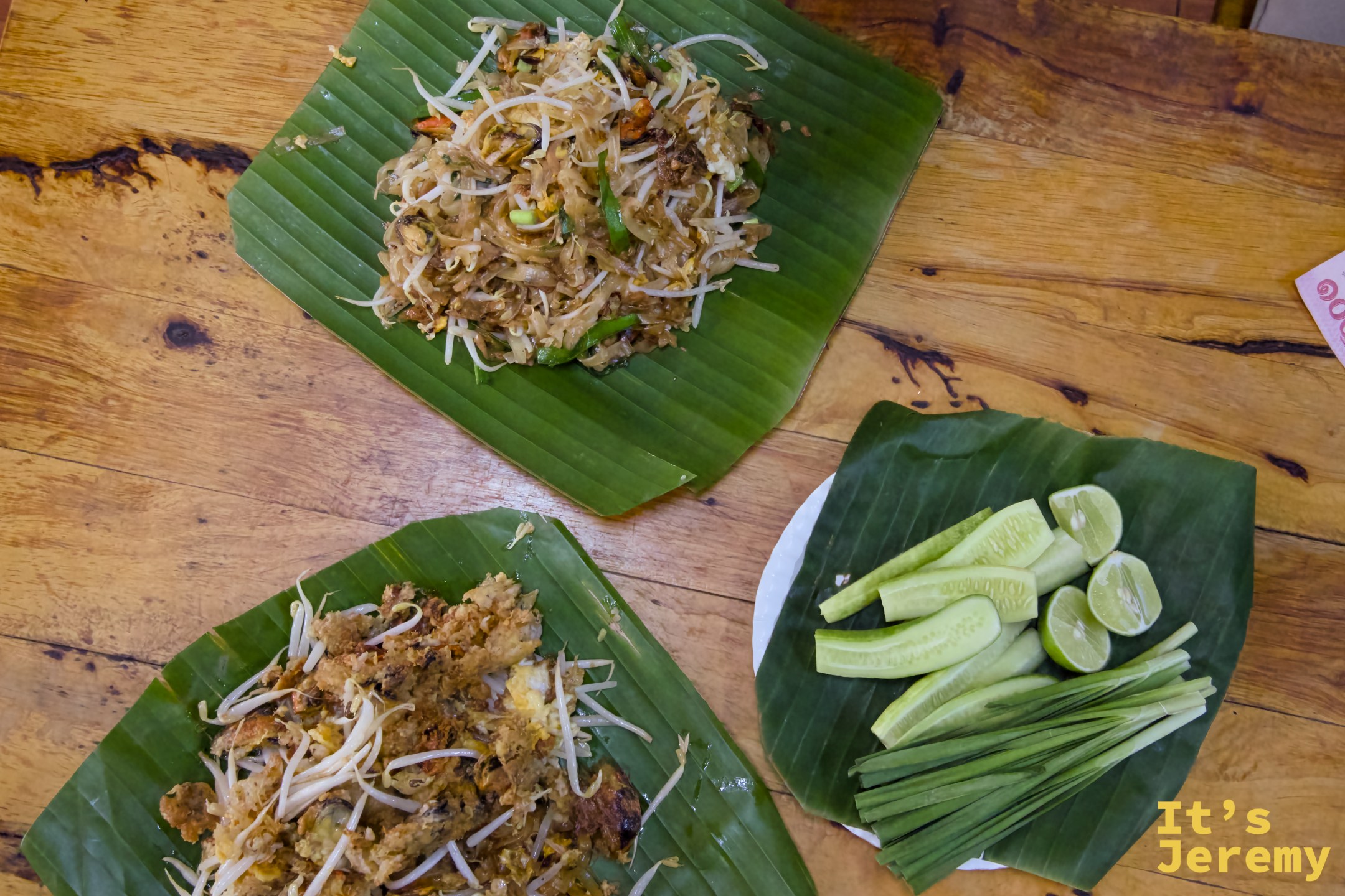 桌面鳥瞰：上方一份 Pad Thai、下方一份 Fried Mussel，右邊是配菜盤（小黃瓜、韭菜、萊姆），三樣全放在芭蕉葉上