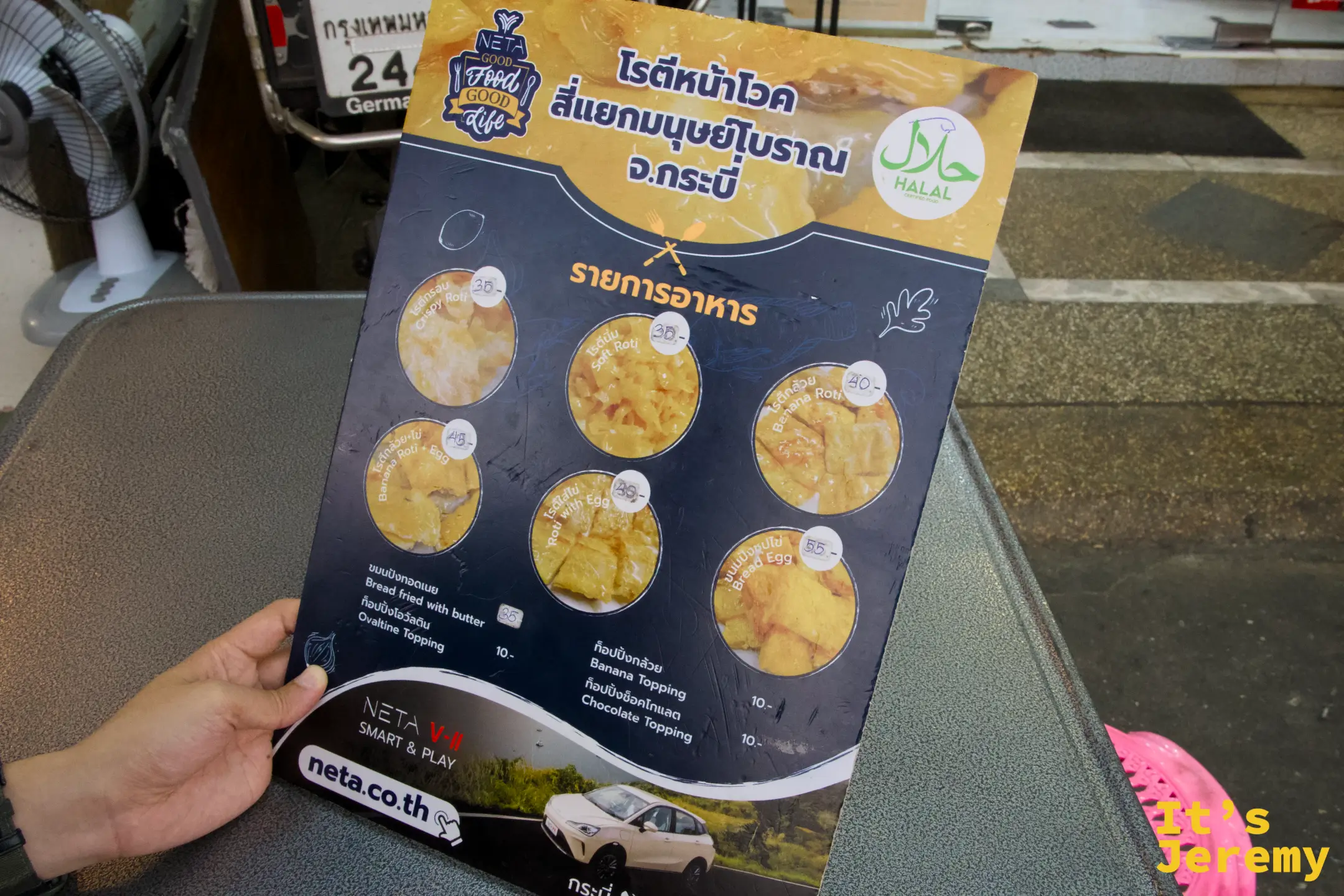 手持菜單，清楚列出各款 Roti 選項、Halal 認證標誌，以及 NETA 汽車廣告背面——典型喀比路邊攤的視覺混搭。