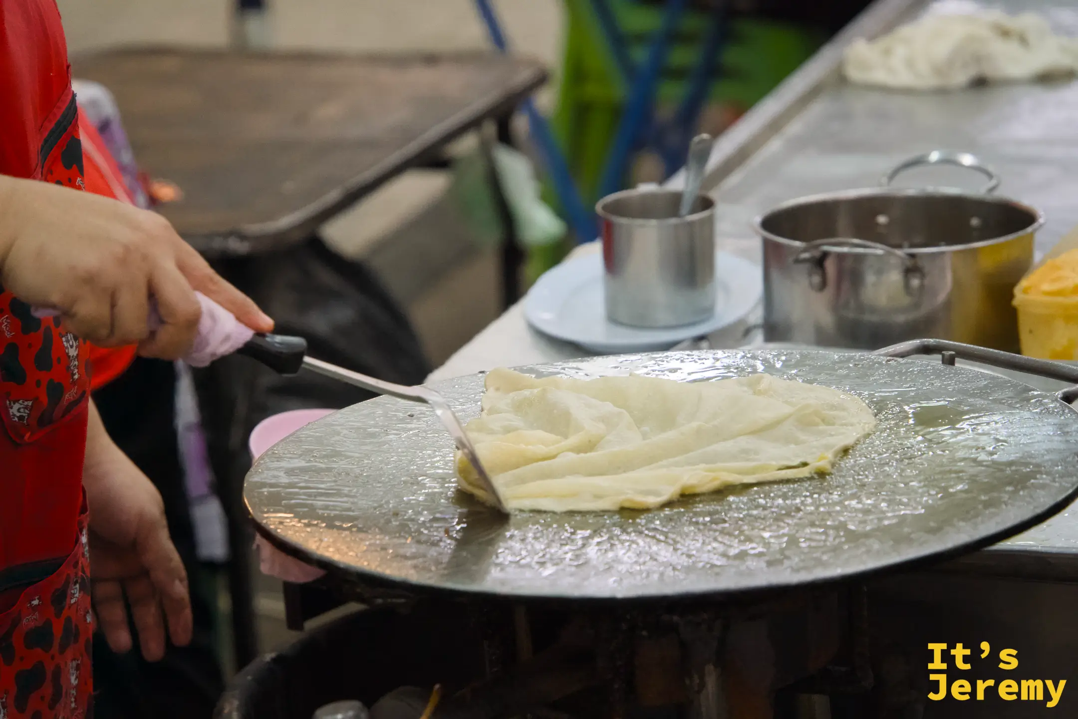 師傅在大圓鐵板上煎著一張薄透的千層 Roti 麵皮，麵皮邊緣呈現焦黃色。