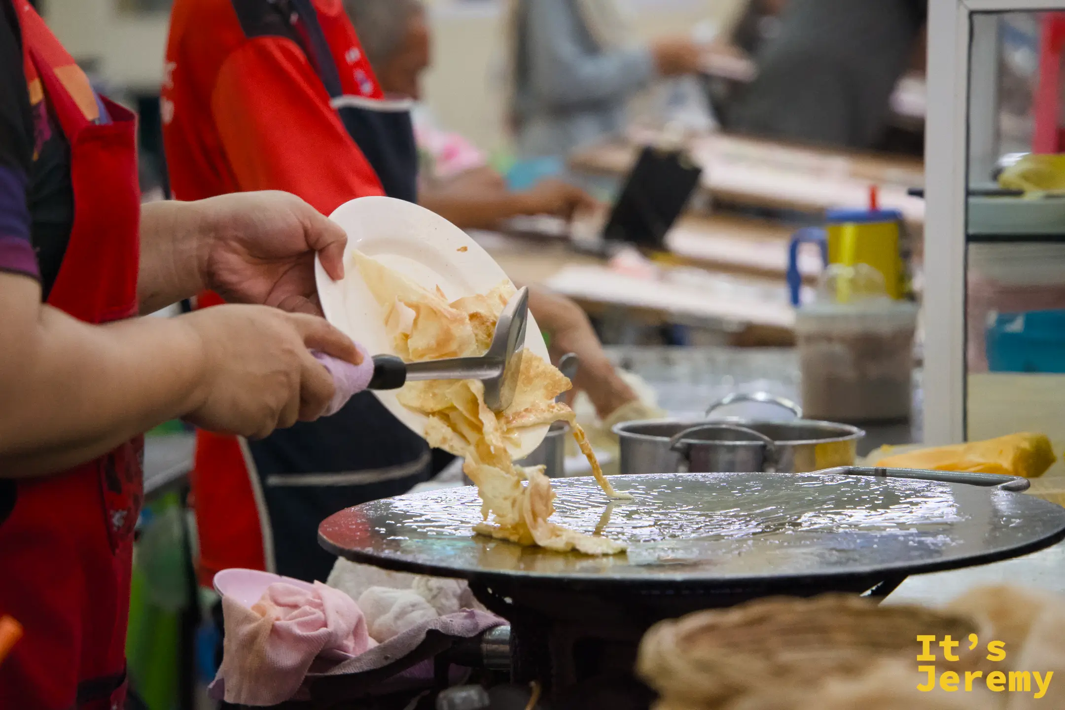 師傅將煎好的千層 Roti 用鏟子鏟起放入盤中，飛濺的油光在盤邊。