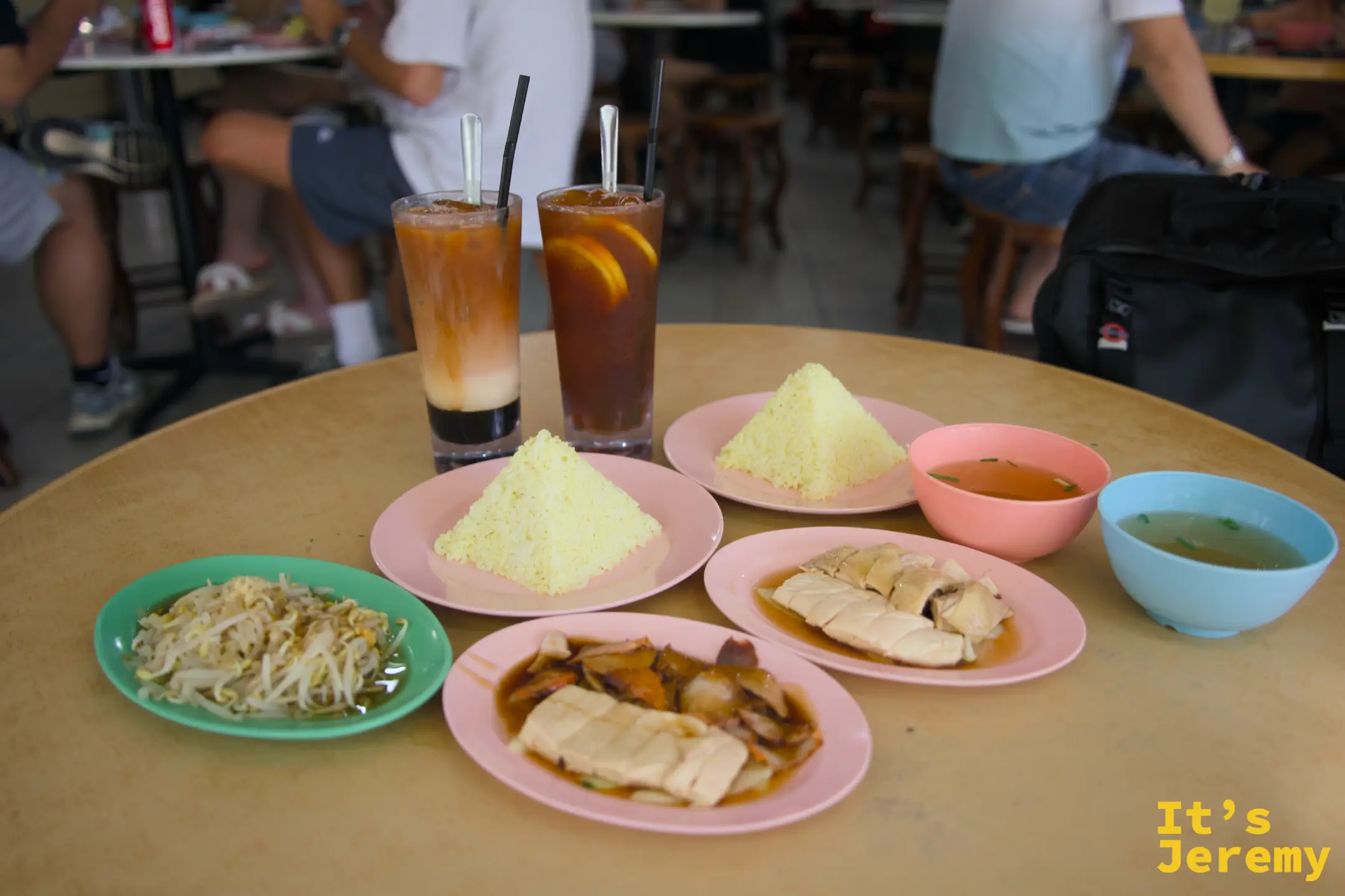 正面視角看全桌：後排兩杯高腳飲料各插吸管，前排兩份金字塔飯、叉燒盤、白切雞盤，左側綠盤芽菜，兩側各有一只湯碗