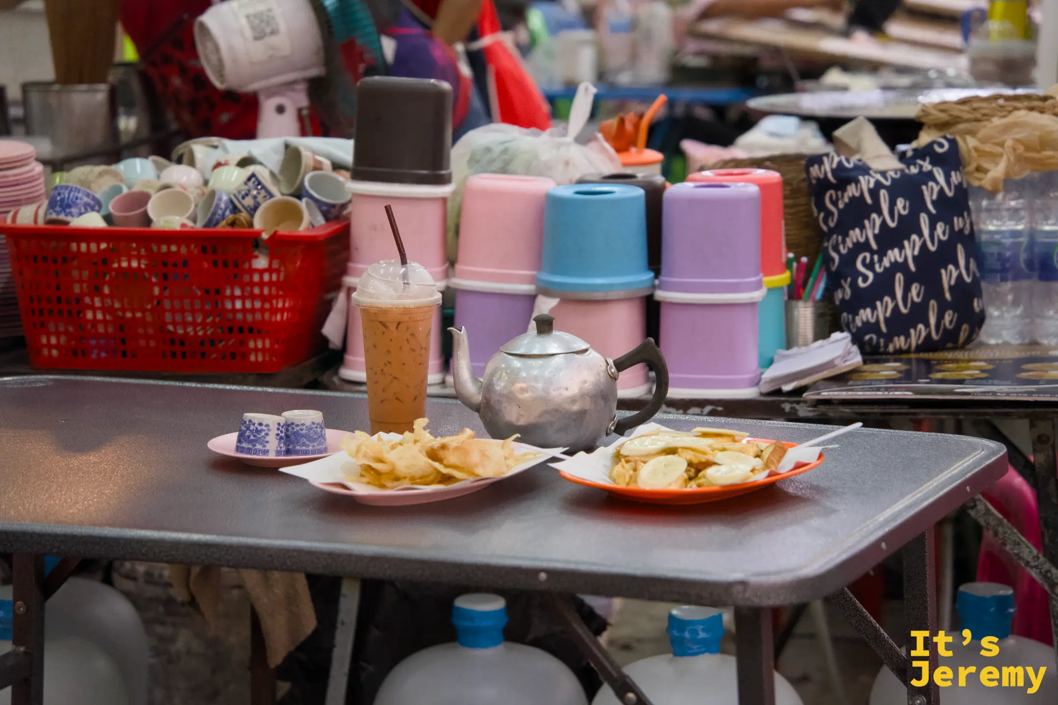 攤前桌面上擺著兩盤 Roti 點心、一壺鐵製熱茶壺和一杯冰奶茶，背景是疊放整齊的各色塑料杯與茶具籃。