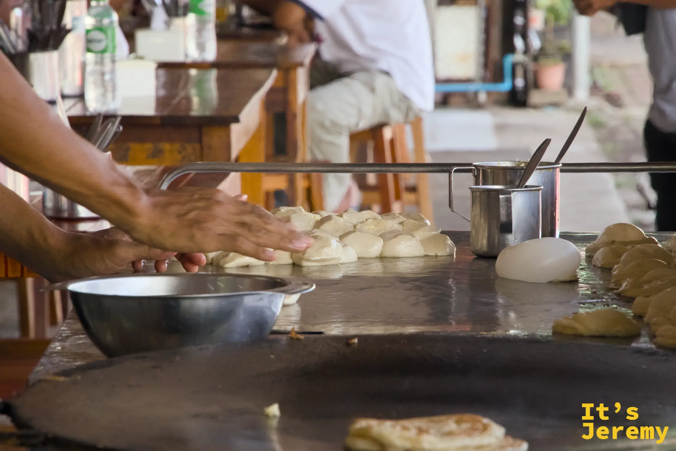 雙手將麵糰鋪展在鐵板上，右側的圓鐵盤上已有一張 Roti 開始煎制