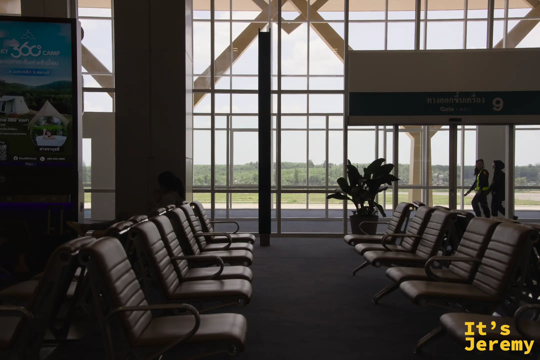 喀比機場候機室,Gate 9 登機門,大片玻璃窗正對停機坪與遠方綠意,兩名地勤人員走過