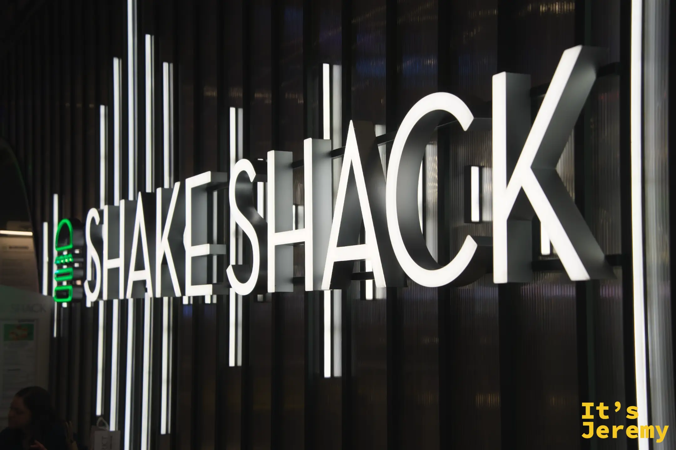 Shake Shack 發光招牌特寫，白色大字在黑色縱條格柵牆上，左側綠色漢堡 logo