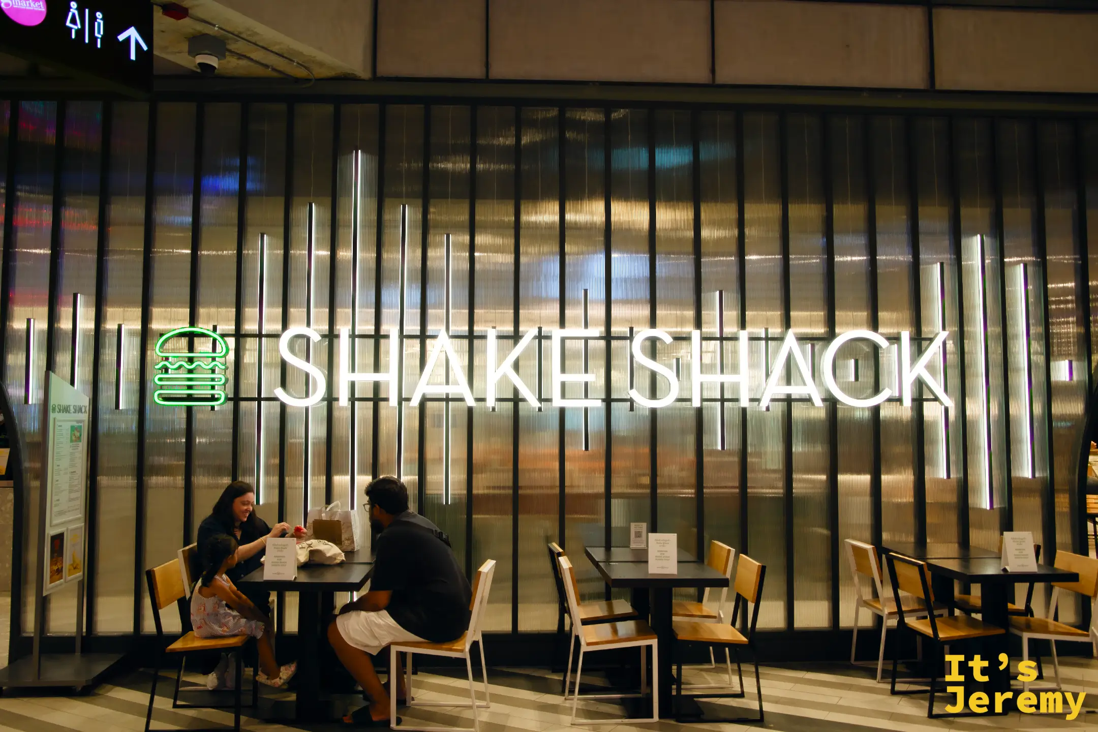 Shake Shack Emsphere 店面全貌，招牌打亮，顧客坐在店外木椅桌用餐，上方 Gourmet Market 指示牌