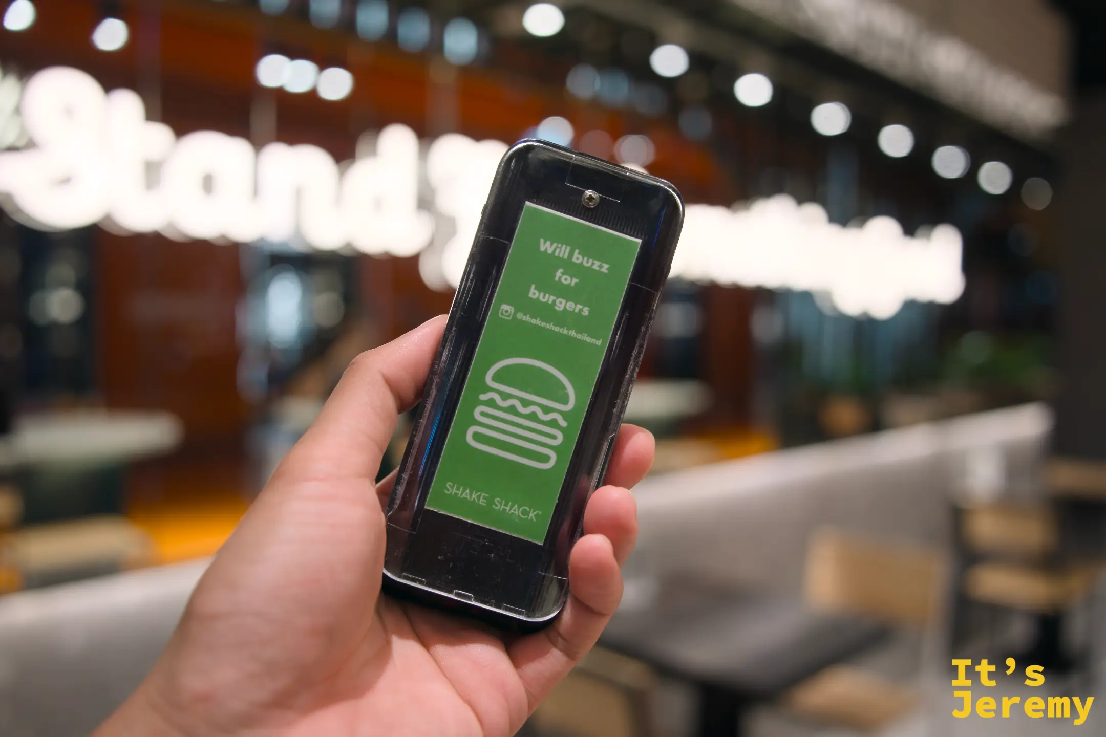 手持 Shake Shack 取餐震動器，正面印「Will buzz for burgers」和漢堡圖案，背景是店內空間