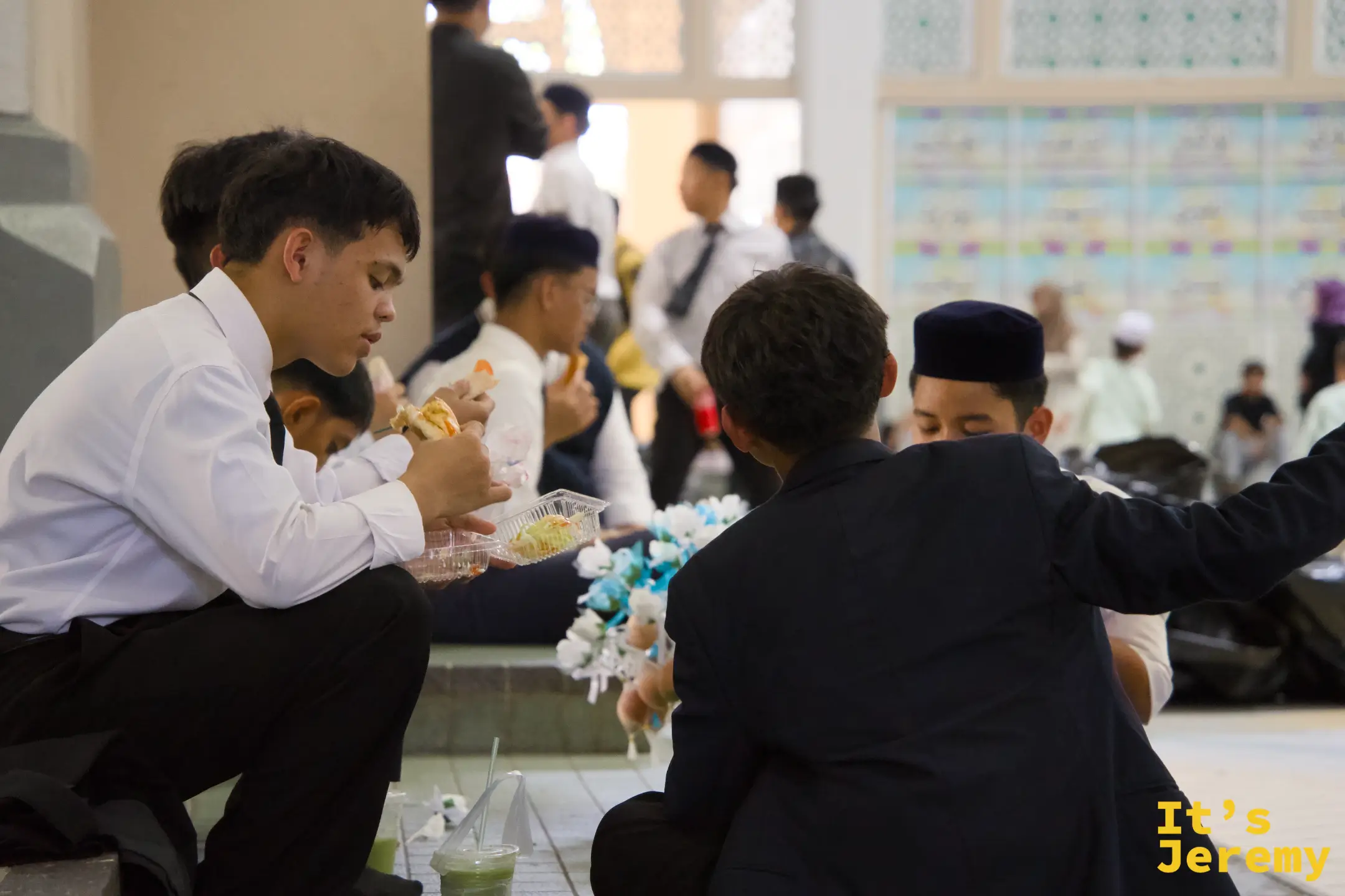 穿著正式西裝的婚禮賓客在廳外聚集，手持 bunga telur 與外帶食品