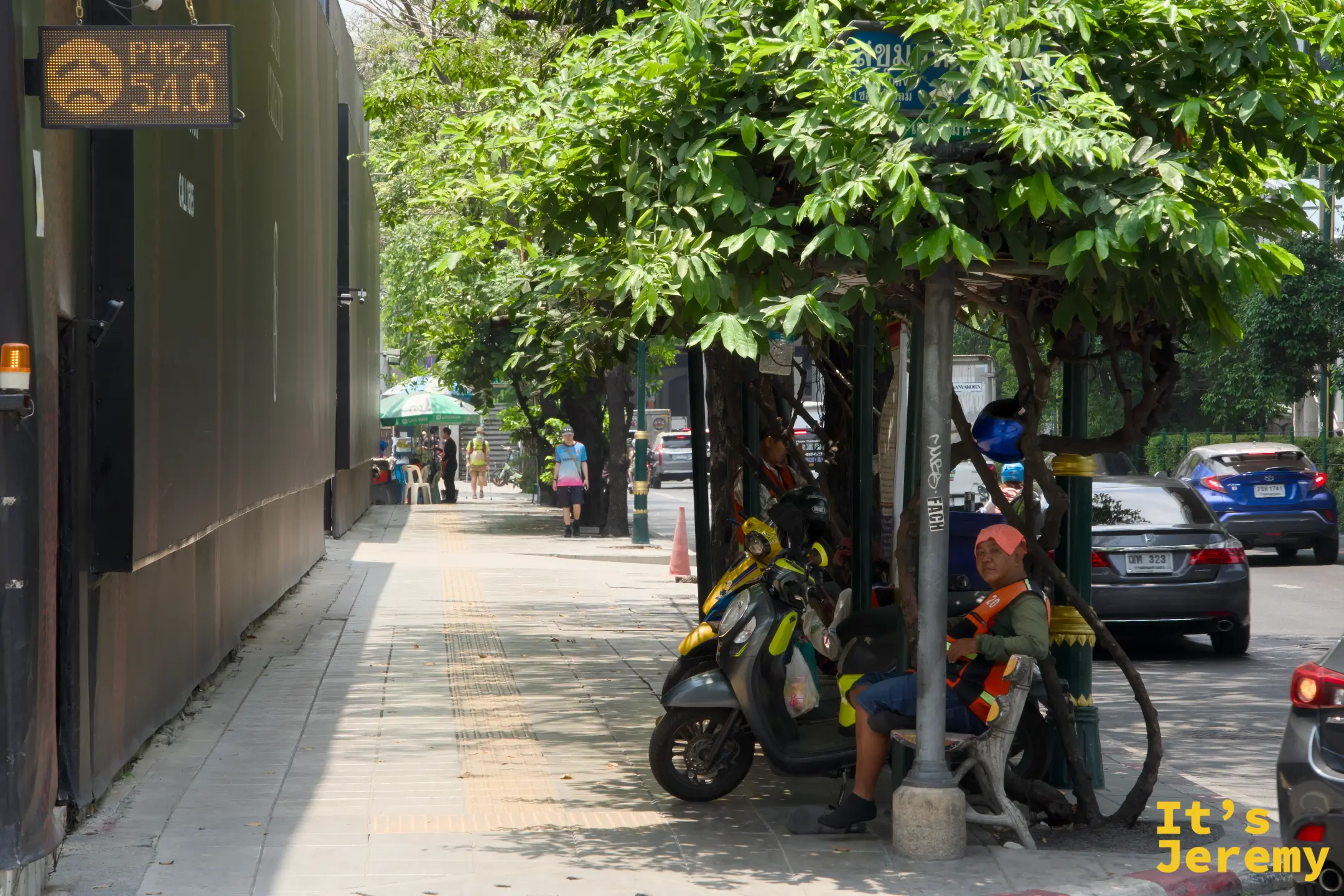 路旁建築牆上的另一塊 PM2.5 電子看板顯示數值 54.0,下方可見幾個工人在僅有的樹蔭下納涼