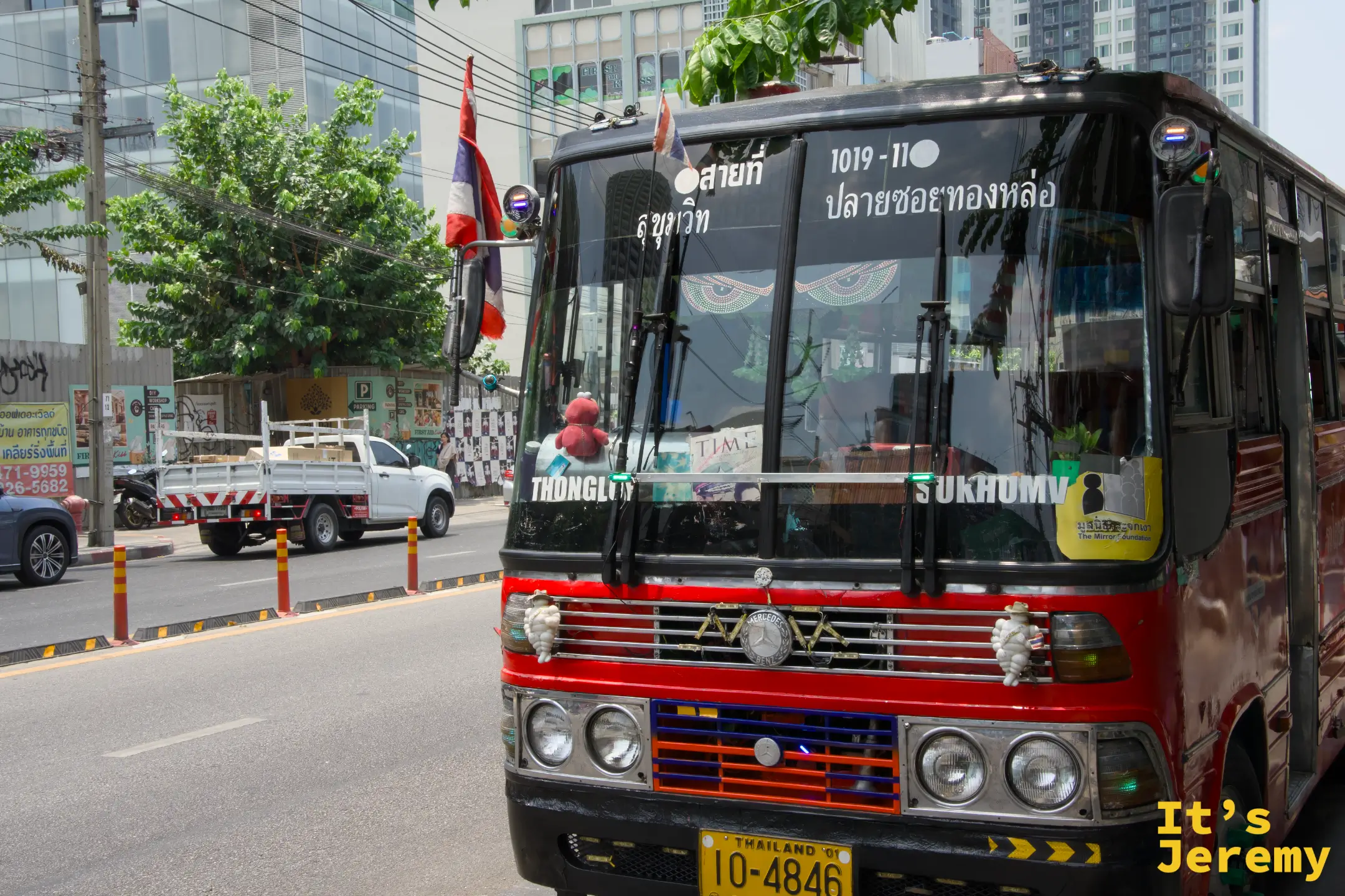 一輛紅黑色的曼谷老公車停在路旁,車身已有些年紀,和不遠處的 BTS 高架橋形成有趣的時代反差