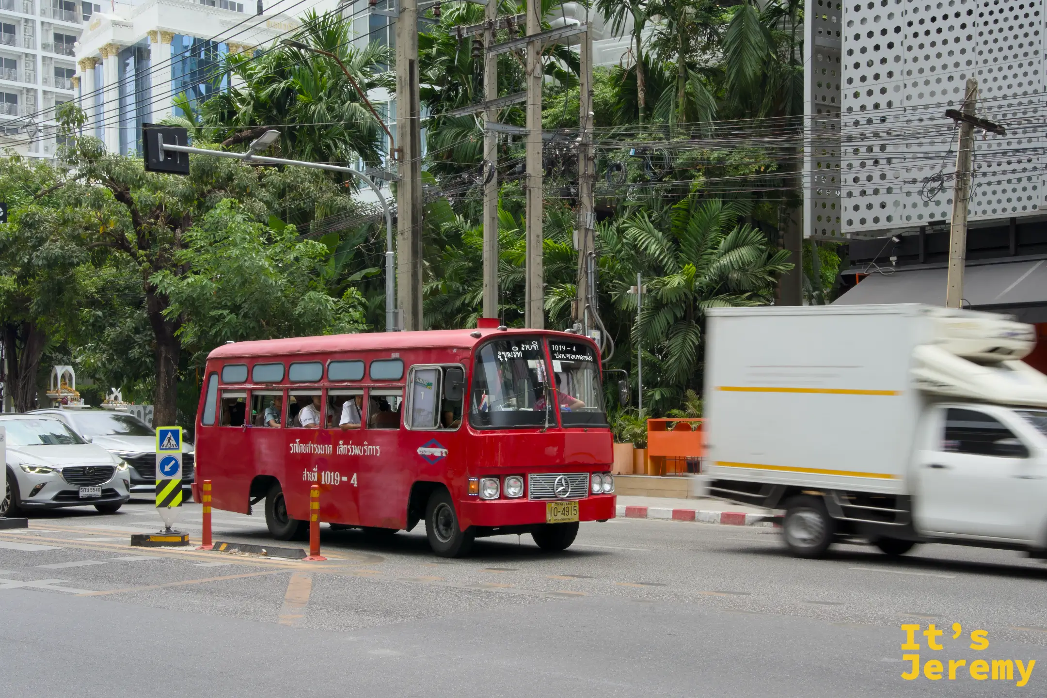 一輛小型紅色曼谷公車穿過路口,舊款車身的顏色在陽光裡顯得有點褪,走進熱氣中