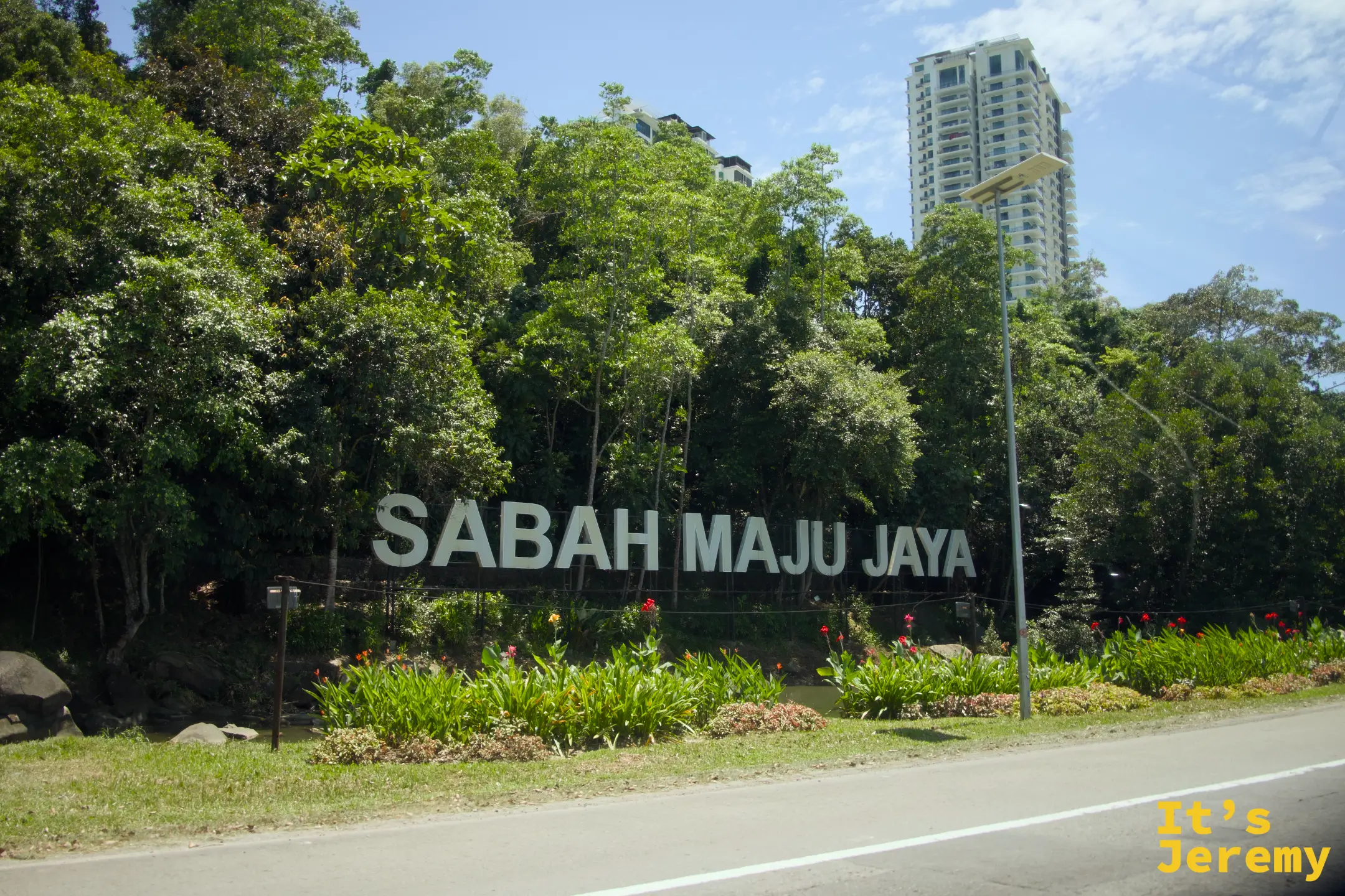 路邊「SABAH MAJU JAYA」大型標牌，綠樹掩映，後方可見高樓