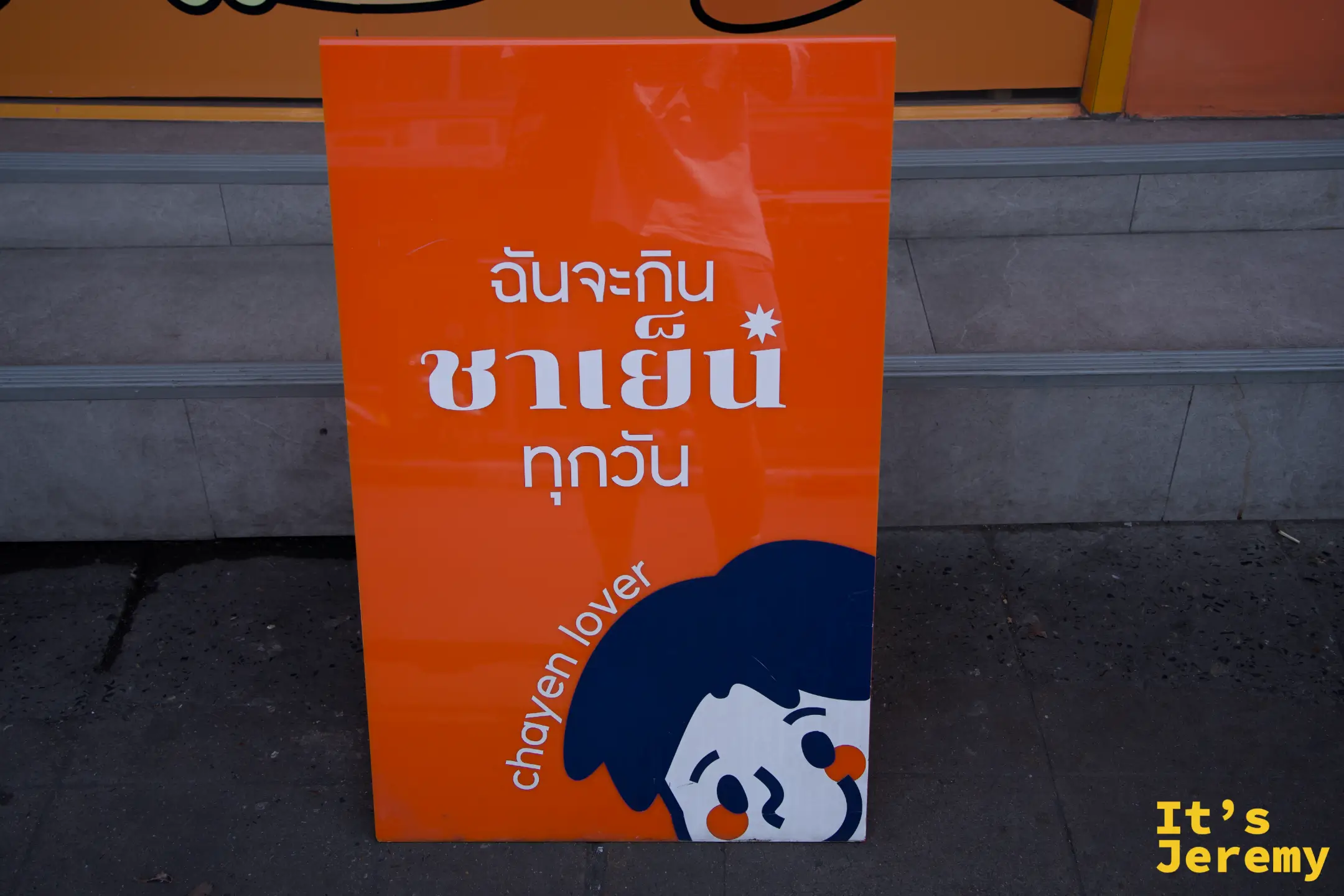 橘底白字的 A 字板，寫著「ฉันจะกิน ชาเย็น ทุกวัน」，chayen lover 角色從下方探出頭來
